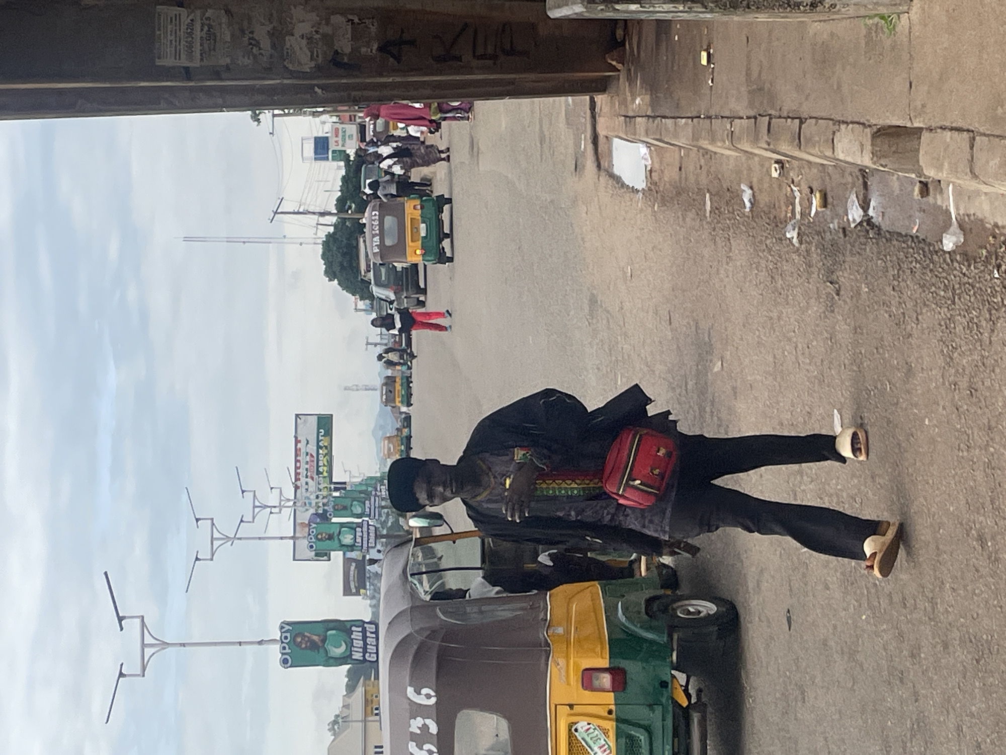 The image captures a street scene in Gura Top, Nigeria. A man, possibly of Nigerian descent, walks towards the camera on the right side of the street. He is wearing a black jacket over a patterned shirt and black trousers, with white sandals on his feet. A red sling bag is worn across his chest. To his left, a yellow and green auto-rickshaw is parked. The street is lined with traffic lights and billboards advertising various services, including "OPay Night Guard." Other vehicles, including more auto-rickshaws and cars, are visible in the background, along with several pedestrians. The environment appears to be an urban setting with a cloudy sky overhead. Debris and water are present on the side of the road.