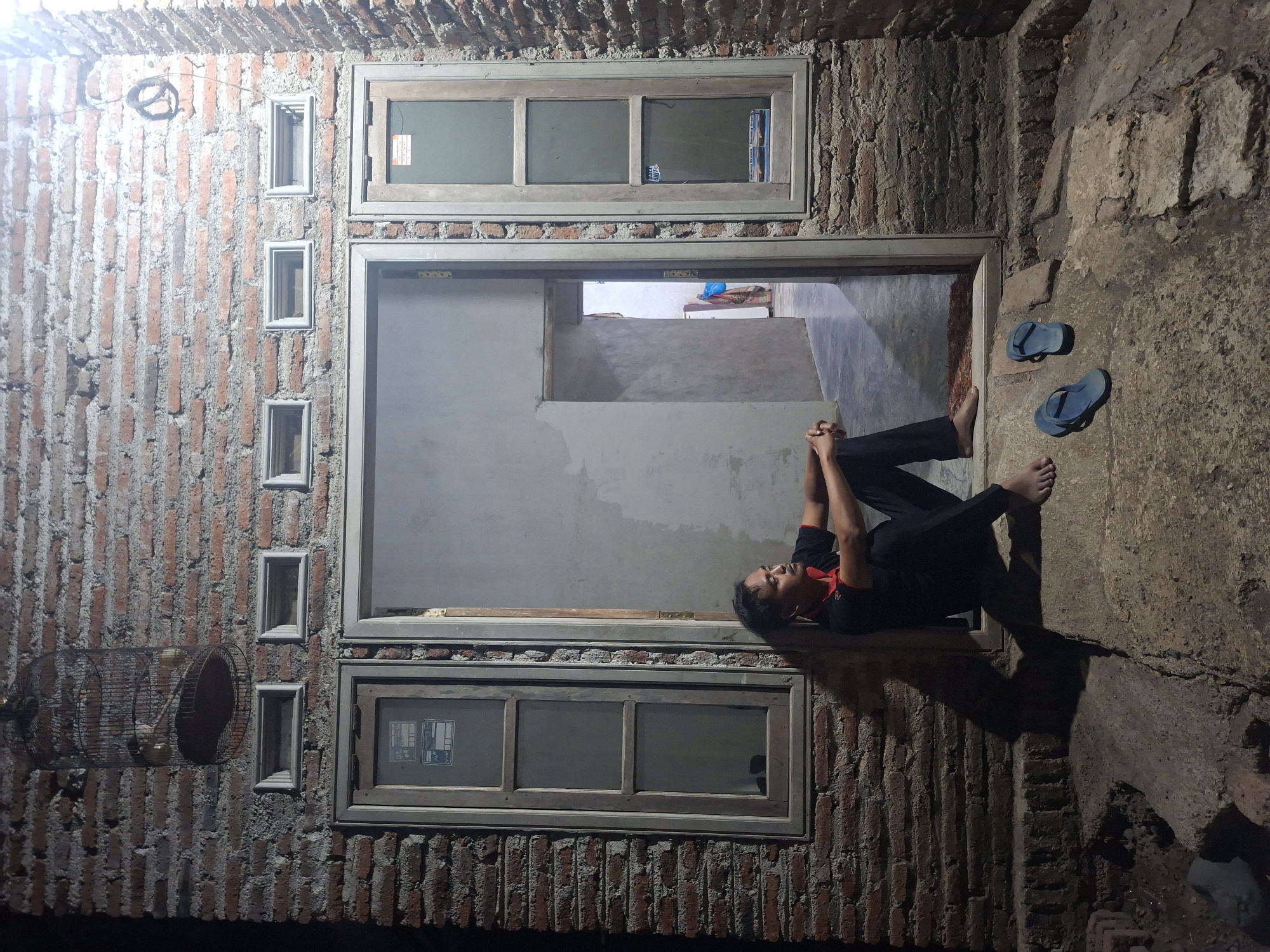A man in a black shirt with a red collar and black pants sits with his legs crossed on a concrete step outside a brick building at night. He is looking to his right. A pair of blue flip-flops are on the ground to his right. In front of him is a doorway leading into a dimly lit interior. To his left is a window with multiple panes. Above the building's brick wall, a birdcage hangs, containing at least one bird. The scene appears to be in the city of Tongaswetan, Indonesia.