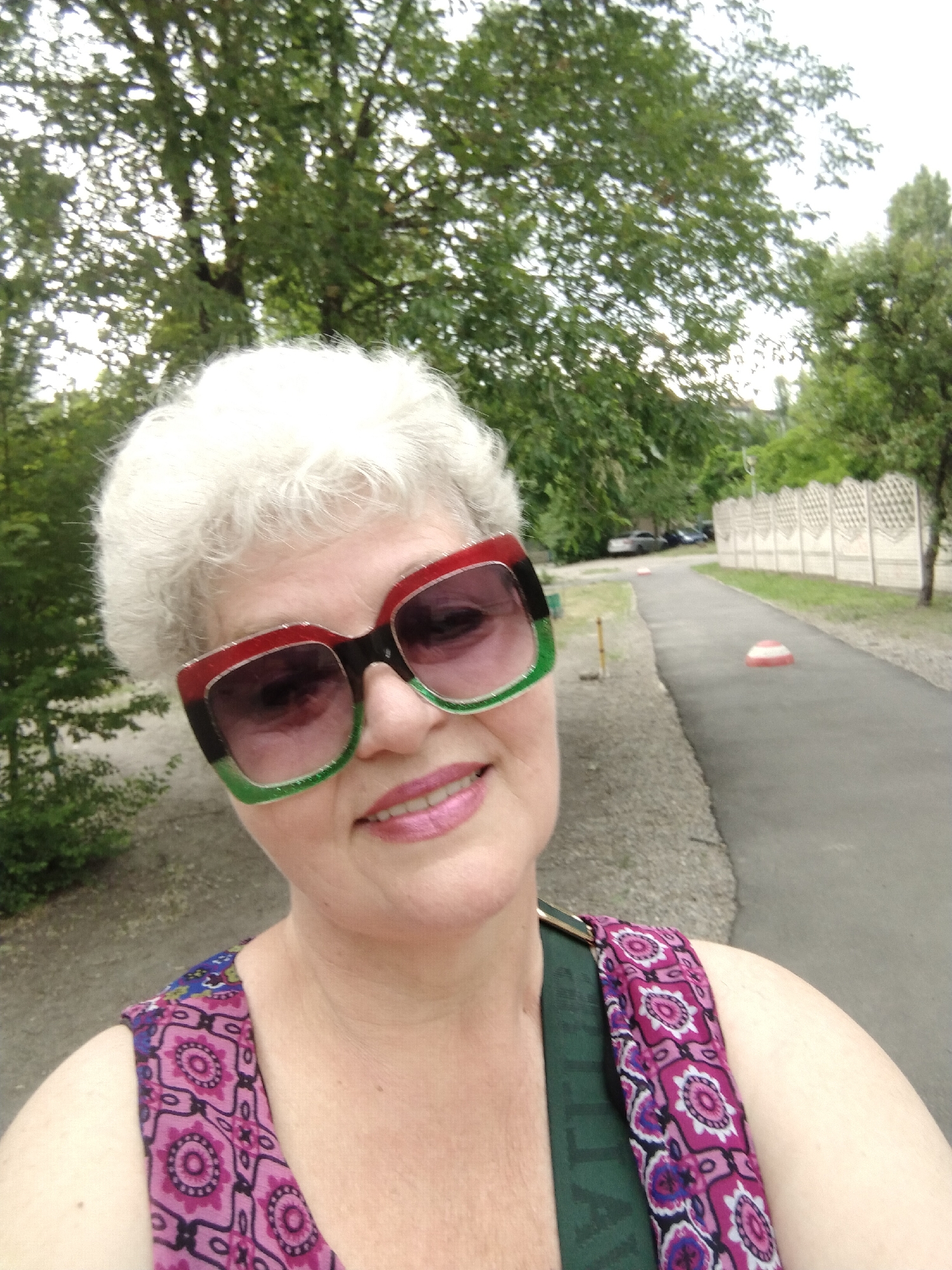 A close-up selfie captures an older woman outdoors on a paved pathway in Horishni Plavni, Ukraine. The woman, positioned in the lower-left and central foreground, has short, curly white hair. She is smiling, revealing her upper teeth, and wears bright pink lipstick. She wears large, rectangular sunglasses with a distinctive tri-color frame: red on top, black in the middle, and green on the bottom, all edged with a thin, glittery silver line. The sunglass lenses are tinted dark purple/brown. Her visible clothing consists of a patterned strap over her left shoulder, featuring fuchsia, black, white, and blue circular motifs on a dark pink background. A dark green strap, likely from a bag, is visible on her left side, partially obscured by her shoulder.
The background consists of a green, arboreal environment. To the immediate left and extending upwards, dense green foliage from trees is visible against a light sky. To the right, a dark gray asphalt pathway recedes into the distance. Alongside the pathway on the right is a light-colored, decorative concrete fence composed of multiple panels. Further down the path, two red and white striped traffic cones are placed on the ground. In the far background, beyond the trees and fence, several parked vehicles and indistinct buildings are faintly visible. The lighting is soft and diffused, suggesting an overcast day or shaded area.