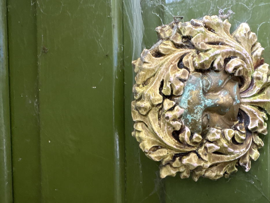 A close-up, vertical shot depicts an aged, ornate metallic decoration mounted on a dark olive-green painted surface, likely a door, in Stiperstones, United Kingdom. The decoration is a relief sculpture of a human face, characteristic of a "Green Man" motif, with foliage surrounding and intertwining with the facial features. The metallic element, appearing to be brass or bronze, exhibits significant verdigris (green patina) on the face and some of the leaf details, alongside areas of original golden-bronze color and darker tarnish. The painted surface behind the ornament shows horizontal panel lines and is covered with extensive, fine cobwebs and scattered dust particles, indicating outdoor exposure and lack of recent maintenance. No people, subjects, actions, or interactions are visible.