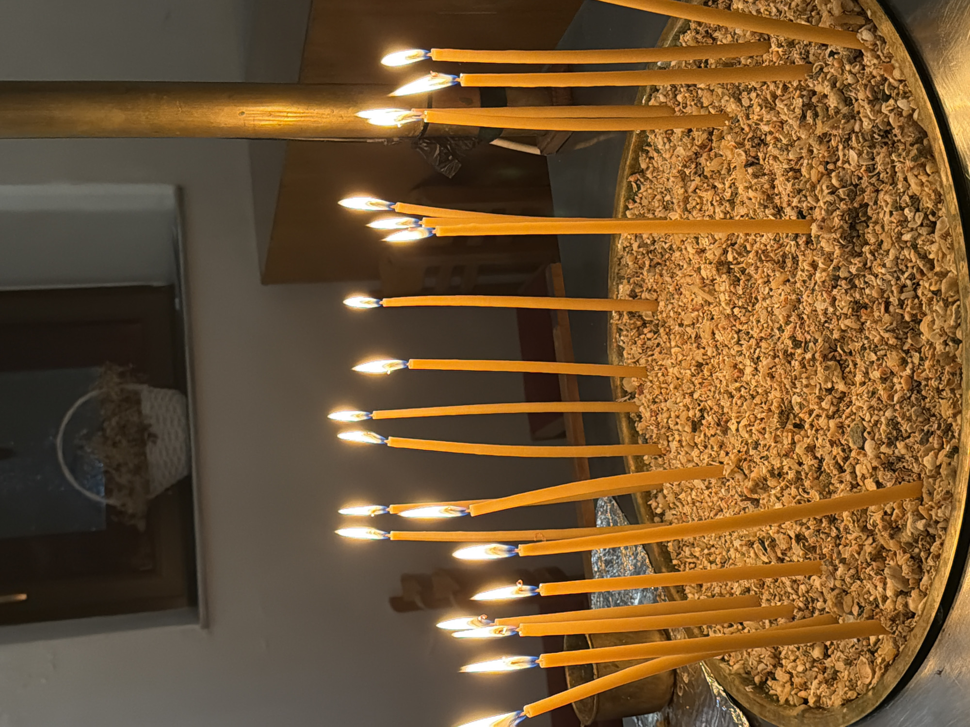In the city of Elafonisos, Greece, a close-up, eye-level shot focuses on a brass censer filled with sand and pebbles, supporting numerous lit, thin yellow taper candles. The candles, varying in height, are arranged in a semi-circular pattern within the censer. Each candle flame flickers, casting a warm glow on the surrounding sand and on the other candles. In the background, a muted, neutral-toned wall is visible, with a dark-framed window featuring a white woven basket holding dried plants. To the right of the window, a cylindrical, metallic fixture, possibly a lamp or part of a support, is present, also catching the ambient light. The scene conveys a sense of quiet contemplation or religious observance within an indoor setting.