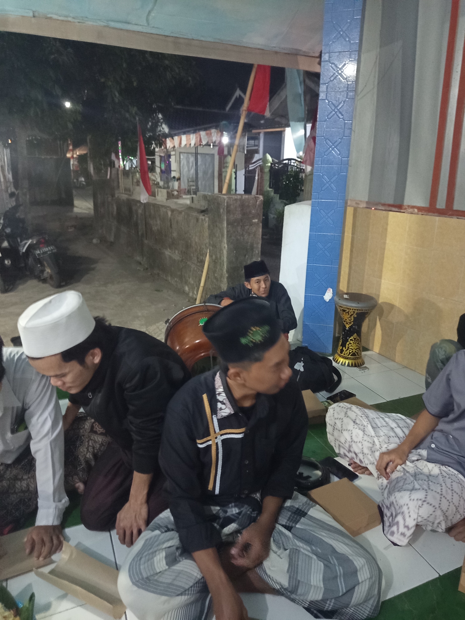 The image depicts an outdoor gathering in the city of Ngablak, Indonesia, at night. Several individuals are seated on the ground, engaged in conversation or attending to something on their laps. One person in the foreground wears a white peci (a cap) and a black robe, while another to his right wears a black peci and a black shirt with white and gold trim. A third person, partially visible on the right, is also seated. In the background, there is a motorcycle parked on a paved surface, and a traditional drum is visible. Flags, including red and white ones, are displayed on poles, suggesting a festive or commemorative occasion. The environment appears to be a street or courtyard, with walls and buildings visible. The lighting suggests it is evening or night.