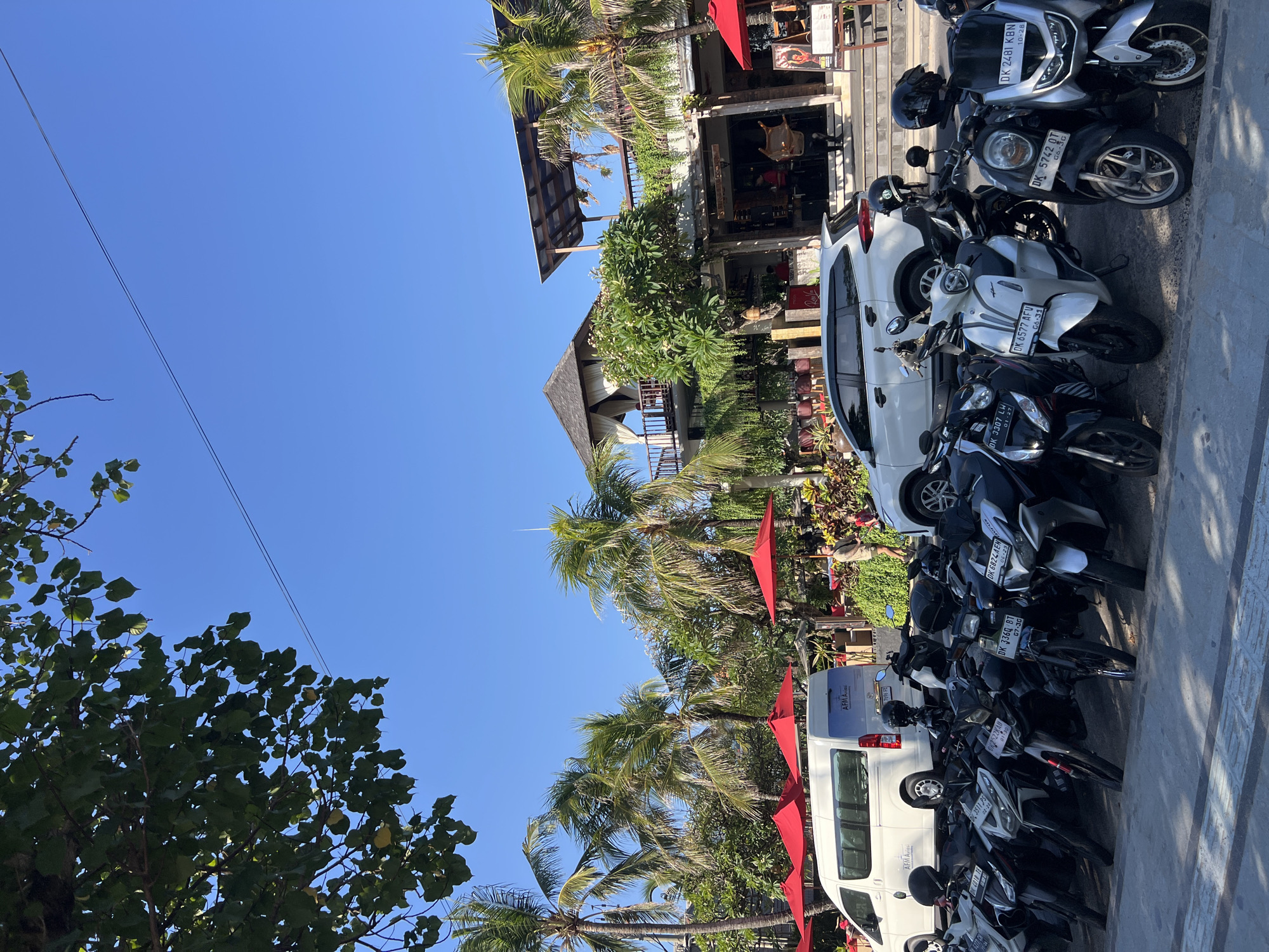 A bright, sunny day in Kuta, Indonesia, is depicted in this vertical, eye-level shot. The scene is dominated by a row of parked motorcycles, with a white van and a white SUV further back. Behind the vehicles, a multi-story building with a distinctive open-air upper level, thatched roof, and balconies is visible. Palm trees and lush greenery surround the building, and several red umbrellas are set up in front of it. The sky is a clear, vibrant blue, with a single power line stretching across it. The street is paved, and the overall impression is one of a tropical, bustling urban environment.