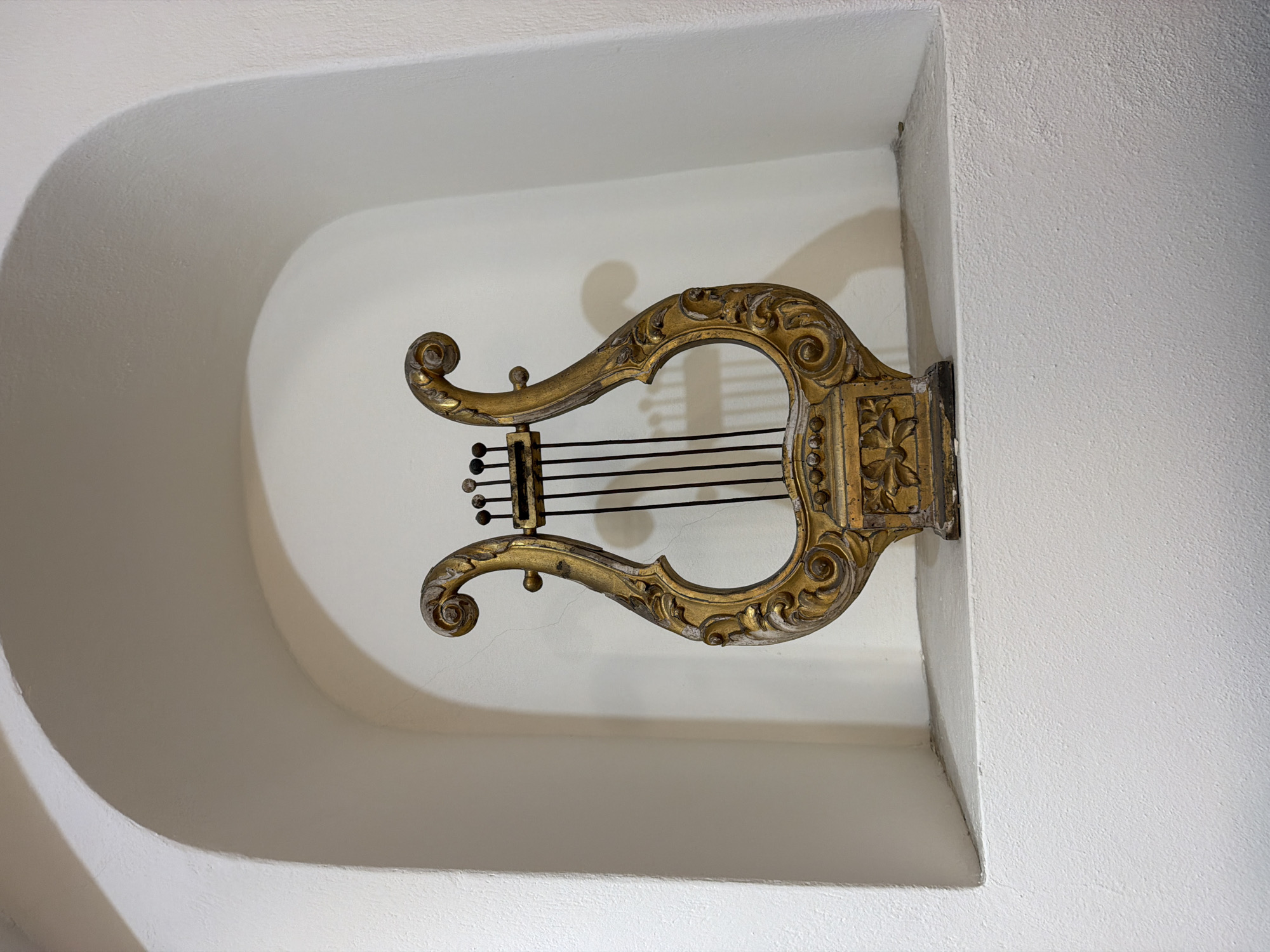 A golden lyre hangs on the wall within a white arched alcove. The strings of the lyre are black. The location is Catania, Italy.