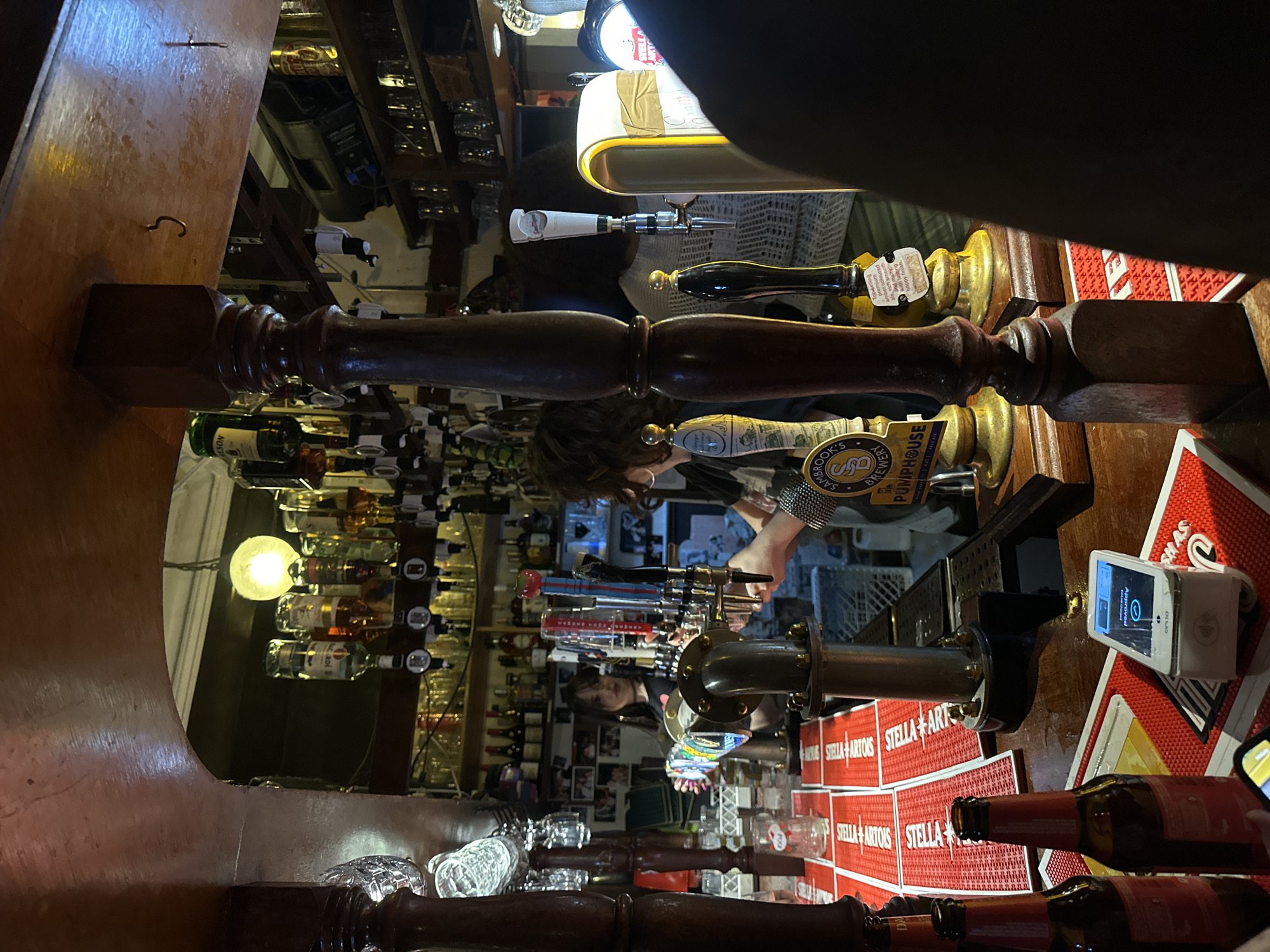 The photograph shows the interior of a bar, possibly in London, United Kingdom. There are several liquor bottles and beer taps behind the bar. The bar itself is made of dark wood. A person is working behind the bar, pouring beer. The image is taken from an unusual perspective, looking up at the bar from a low angle. On the bar top, there are several beer bottles, coasters with the name "Stella Artois," and a card reader. The word "PUMPHOUSE" is visible on one of the beer tap handles.