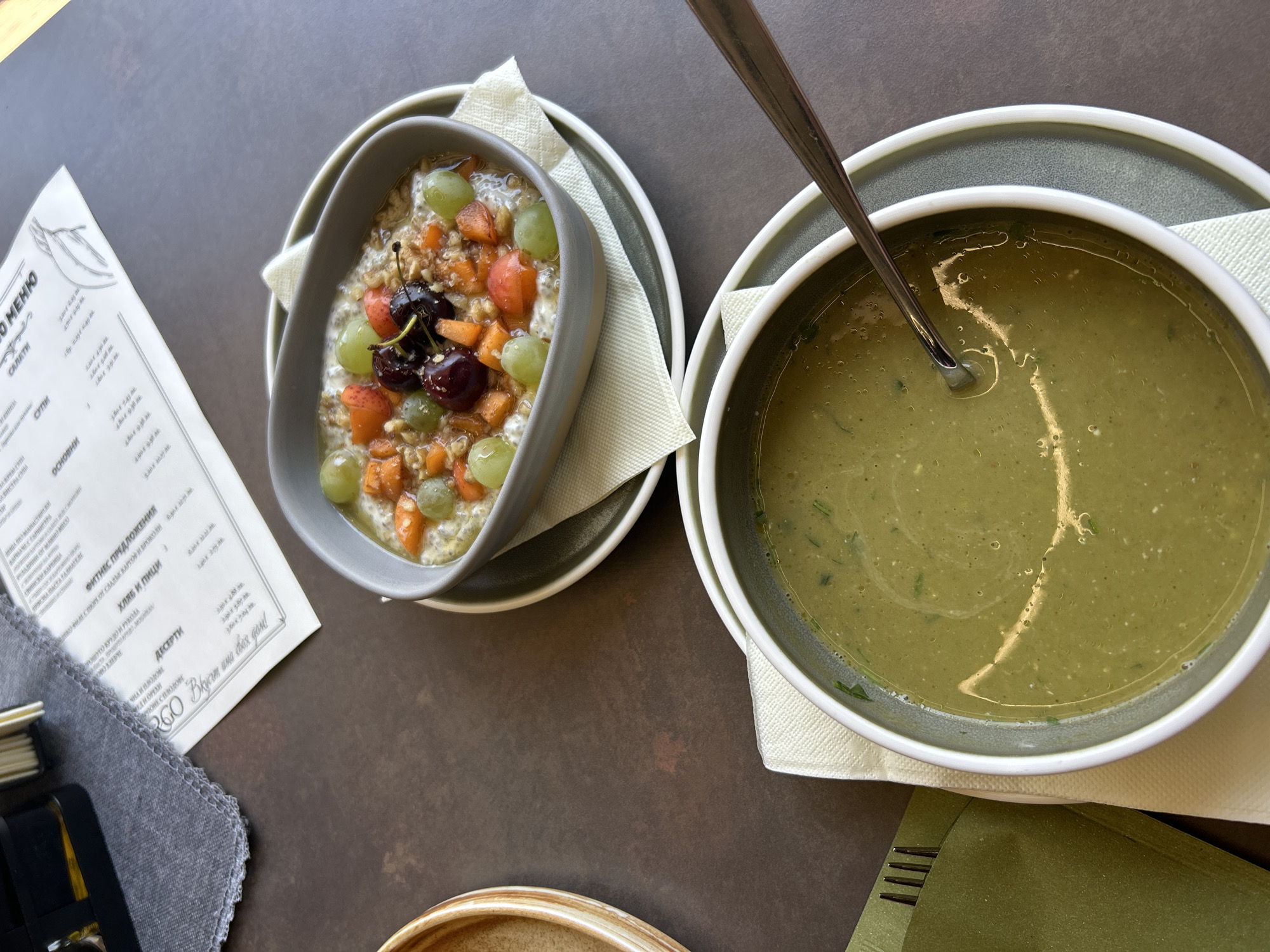This is a top-down, close-up shot of a meal served on a dark, textured table. Two bowls are prominently featured. The bowl on the left contains a breakfast dish, likely oatmeal, topped with chopped fruits like grapes, cherries, and possibly strawberries, along with nuts and seeds. The bowl on the right holds a thick, green soup, with a spoon resting inside. Both bowls are placed on white napkins and gray plates.
To the left of the bowls, a menu is partially visible, printed on white paper with a sketch of a leafy branch. The menu is open, revealing sections labeled "МЕНЮ" (Menu), "САЛАТИ" (Salads), "СУПИ" (Soups), and "ОСНОВНИ" (Main Courses), along with some item names and prices in Cyrillic script. There's also a section for "ФИТНЕС ПРЕДЛОЖЕНИЯ" (Fitness Offers) and "ХЛЯБ И ПИЦИ" (Bread and Pizza), and "ДЕСЕРТИ" (Desserts). The text "Вкусни дни" (Delicious days) is visible at the bottom of the menu. A stack of folded gray napkins or cloths is also on the left side of the image.
The lighting suggests an indoor setting, possibly a restaurant or cafe, with soft, diffused light. There are no people visible in the frame. The overall impression is one of a healthy and appetizing meal, presented in a clean and modern setting.