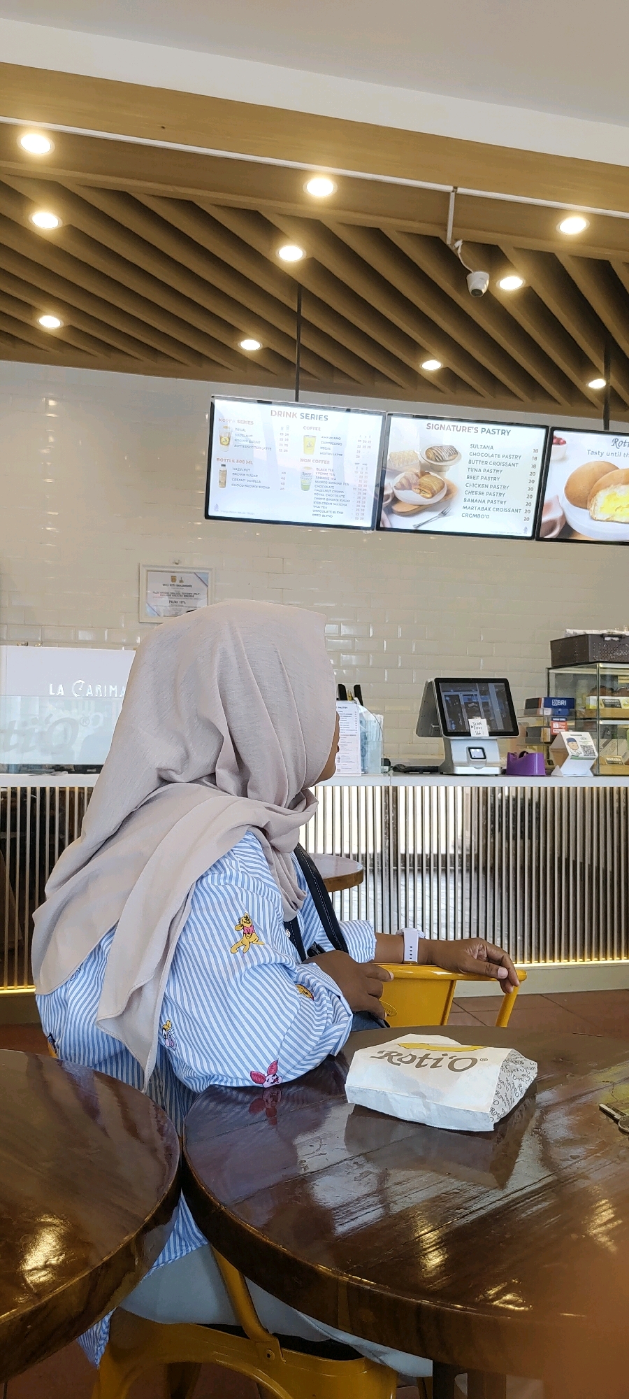 In Banjarbaru, Indonesia, a woman wearing a light brown hijab and a blue and white striped shirt with cartoon characters is seated at a wooden table. She is facing away from the camera, looking towards a counter area. On the table in front of her is a white paper bag with "Roti O" printed on it. The background shows a cafe interior with a menu board displaying drinks and pastries, a cash register, and decorative wooden paneling on the ceiling. The overall impression is a casual dining or cafe setting.