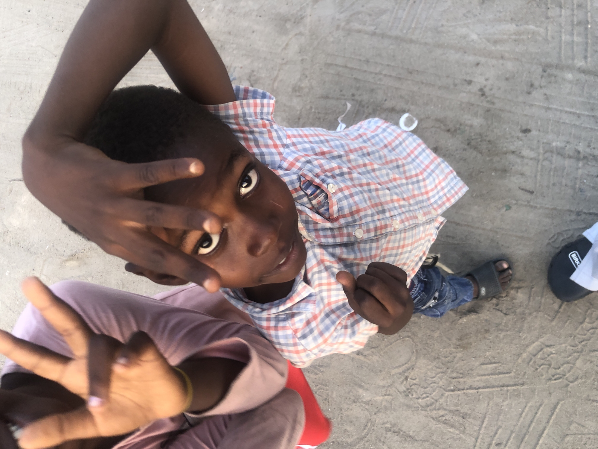 This image, taken in Ado, Nigeria, is a high-angle shot of two young individuals. In the foreground, a boy in a blue and red plaid shirt and blue jeans is looking up at the camera, with his right hand covering part of his forehead and his left hand extended upwards in a peace sign. His left arm is bent behind his head. To the left of the boy, a person wearing a light pink garment is visible, with their left hand raised towards the camera, also making a peace sign. The ground beneath them appears to be a dusty, concrete surface with tire tracks. A red shoe is also visible to the left of the pink garment.