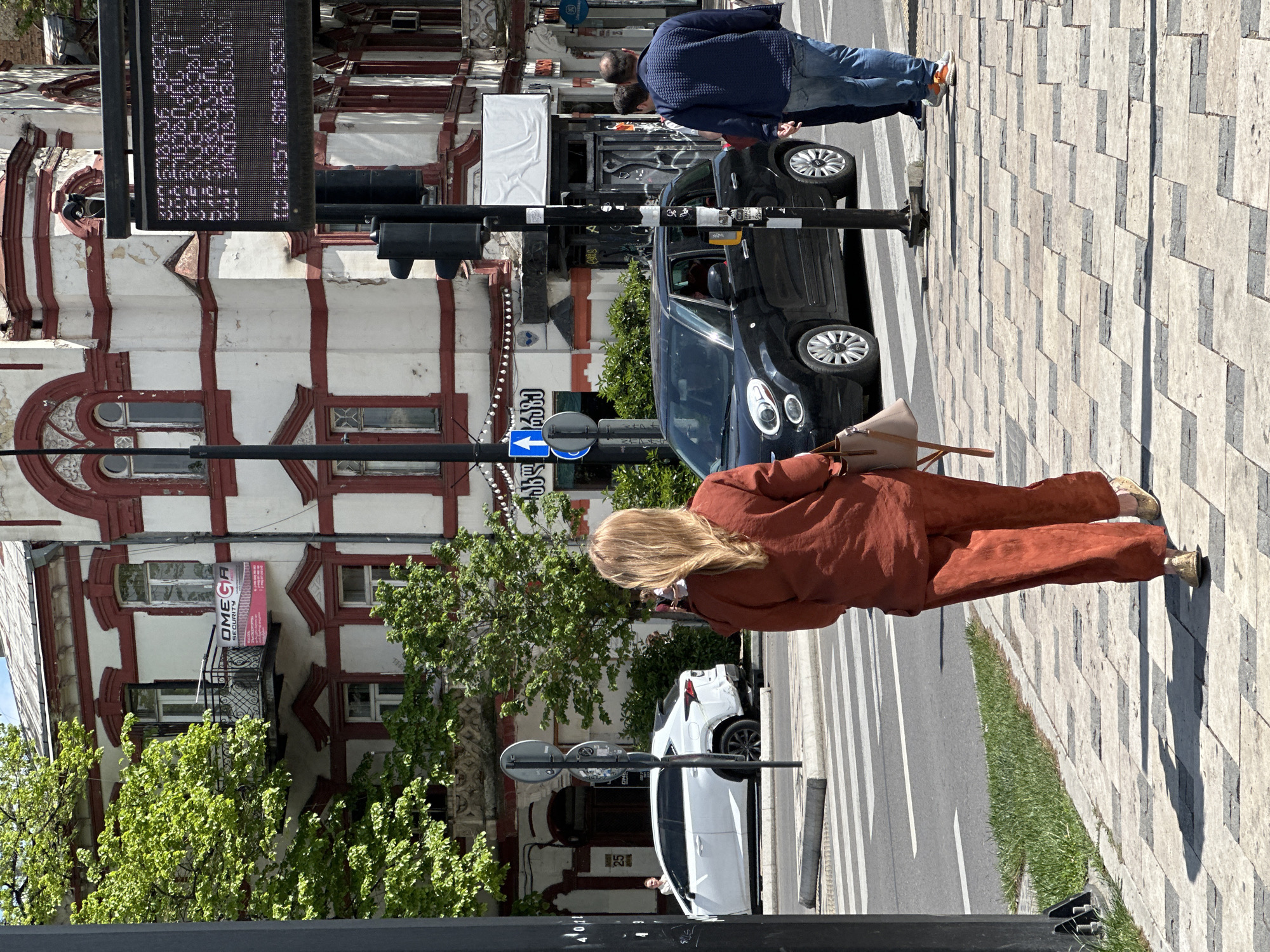 The image depicts a street scene in Tbilisi, Georgia. The shot is taken from an elevated position, possibly a building or structure overlooking the street.
A woman with blonde hair, wearing an orange-colored pantsuit, is walking on the patterned sidewalk. Her back is facing the camera. To the right of her is a narrow strip of grass, then the road. Two cars, one black and one white, are parked along the side of the road.
On the left side of the image is a building with a distinctive white facade and red trim. The building has multiple windows. A sign for "OMEGA SECURITY" is visible on a balcony. There are trees with green leaves in front of the building.
In the foreground, there is a black traffic light pole with a display screen showing text, possibly a news ticker or public information. There are also street signs.
Two men are walking on the sidewalk near the black car. One man is wearing a blue jacket and jeans. The other man is partially obscured behind him.
The scene appears to be a typical day in Tbilisi.