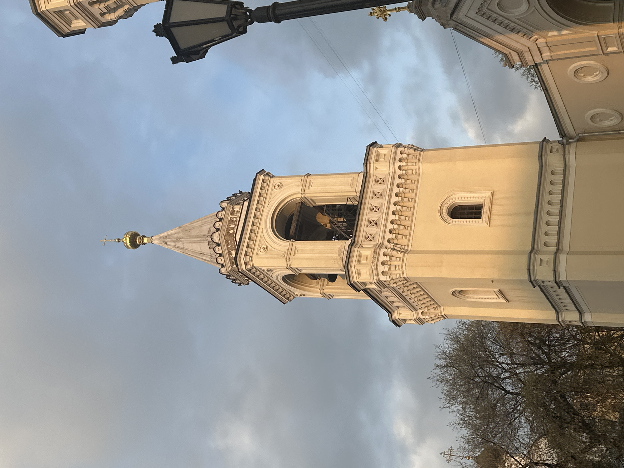 A tall, ornate bell tower of an Orthodox church dominates the central and right portions of the image, bathed in warm, directional sunlight suggesting late afternoon or early evening. The tower is constructed from light cream or pale yellow stone and features intricate architectural details. Its lower visible section includes a small, arched window with a gridded pane. Above this, a prominent tier showcases a large arched opening where bells are faintly visible within the shadow, framed by decorative balustrades, small columns, and patterned relief work. The tower tapers upwards to a slender spire, crowned with a gleaming golden onion dome and a small golden Orthodox cross.
In the upper left corner, an elaborate, dark metal street lamp extends into the frame. Partial views of another building, sharing the same architectural style and color palette as the bell tower, are visible in the upper right. The lower right of the frame is filled with the bare, gnarled branches of a deciduous tree, indicative of winter or early spring. The sky overhead is partially cloudy, with patches of lighter blue sky peeking through. No people are visible, nor is there any discernible text. The scene evokes a sense of historical grandeur typical of religious architecture in Moscow, Russia.
