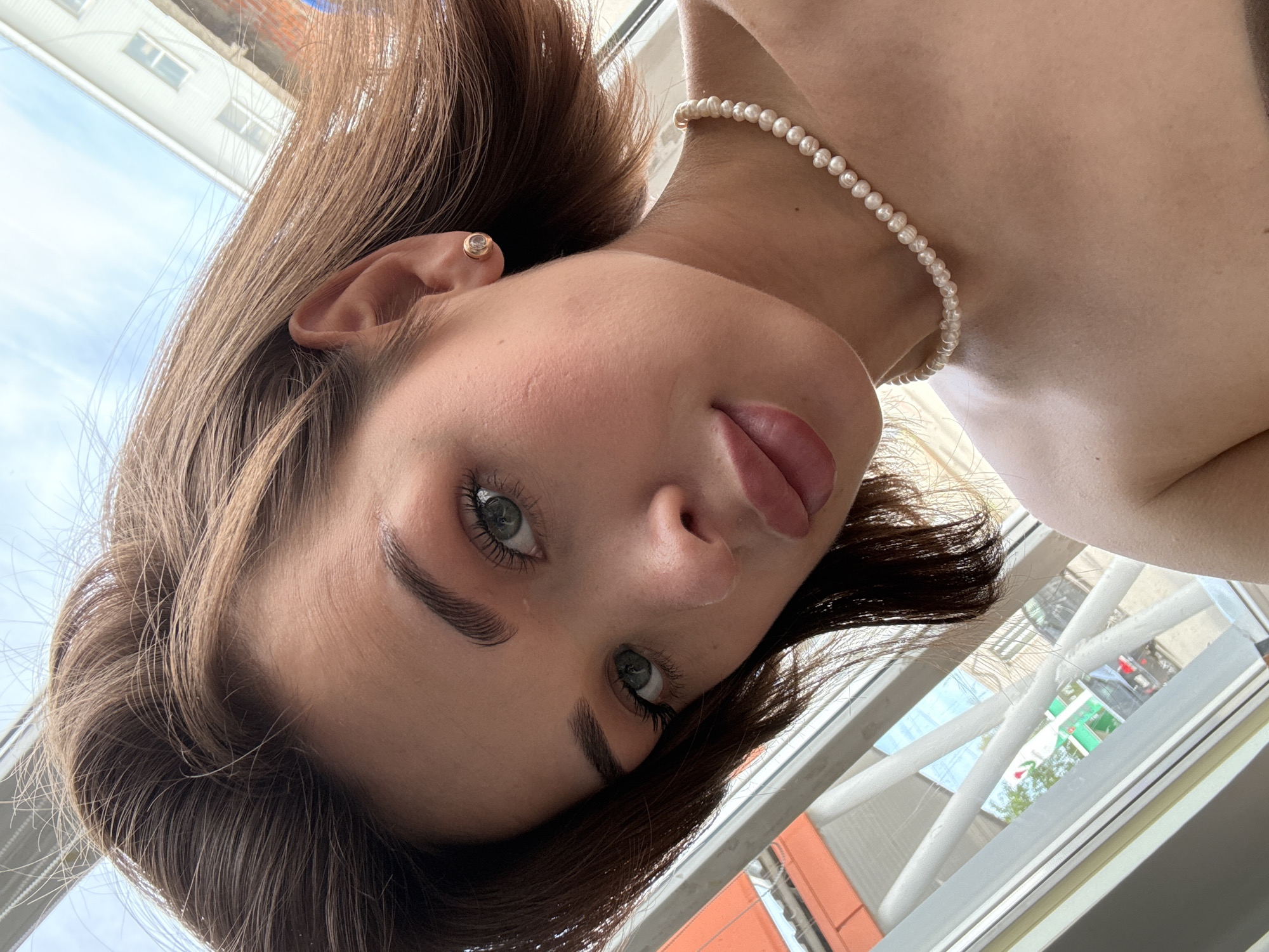 A close-up, angled selfie of a fair-skinned young woman with light brown, shoulder-length hair. She has an earring in her left ear and is wearing a pearl necklace. Her eyes are light colored, possibly green or hazel. She appears to be indoors near a window. Outside the window, visible through the reflections, there are indistinct urban structures. The location in Sarapul, Russia is not identifiable from the image alone.