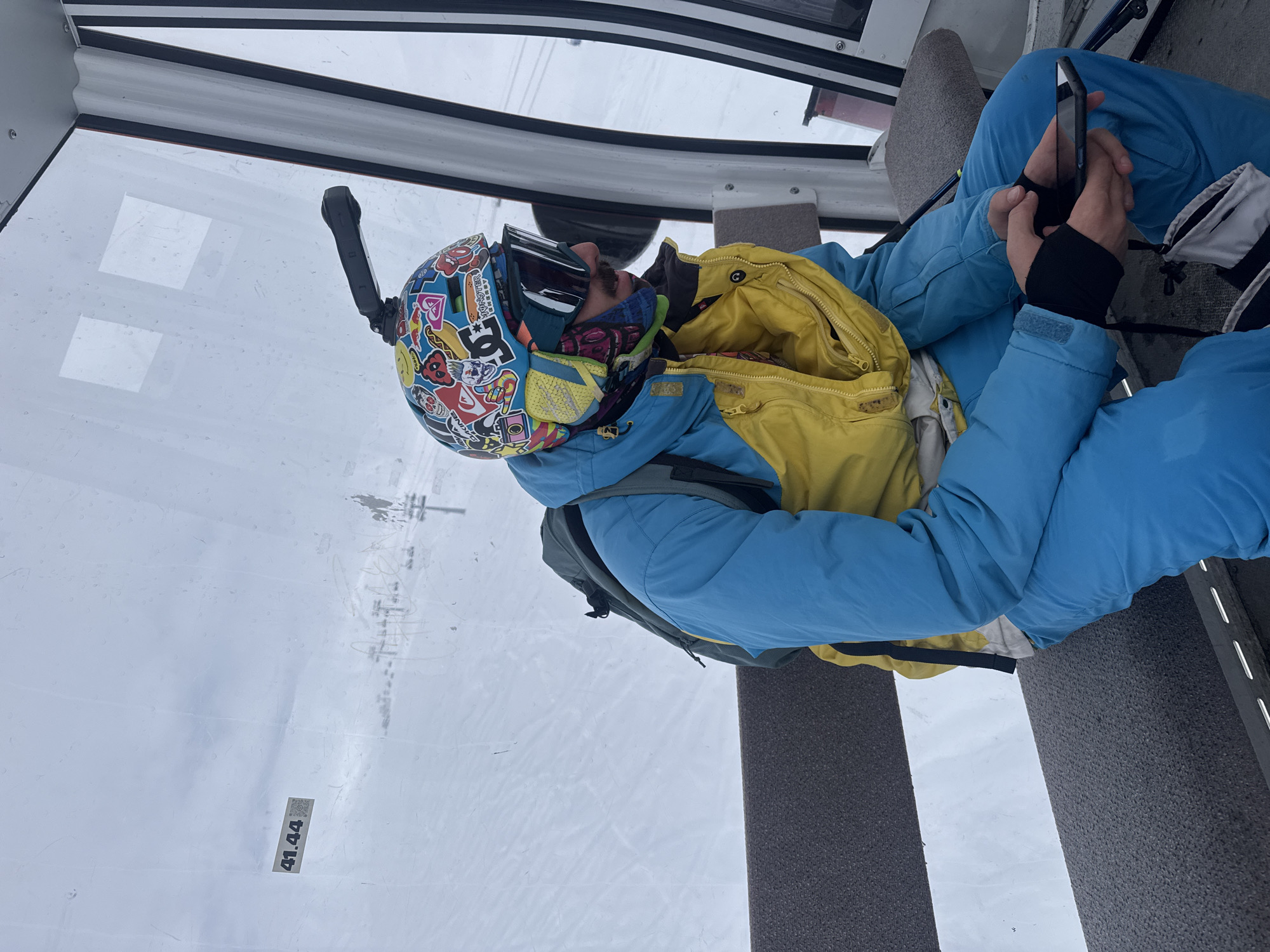 A person is seated inside a cable car or gondola. The individual wears a sticker-covered helmet with an attached action camera, ski goggles, a blue jacket with a yellow interior, and blue pants. They are holding and looking at a smartphone. The cable car has a window with condensation or moisture on the glass, and the scenery outside shows a snowy landscape with a ski lift in the distance. The location is in Unknown, Unknown.