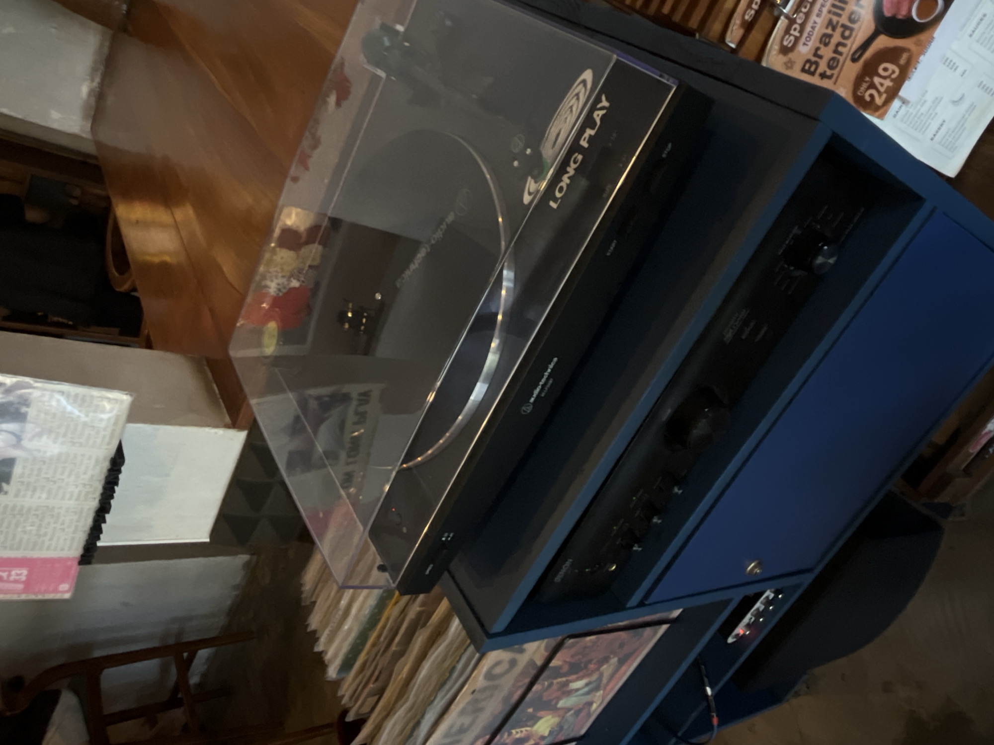 The image depicts a close-up view of a turntable and audio equipment on a blue cabinet, set against a background that suggests an indoor environment with wooden furniture and a wall. The turntable, a black Audio-Technica model, is covered with a clear plastic lid, and the words "LONG PLAY" are visible on the surface. Below the turntable, on the same cabinet, is a black Denon audio receiver. To the left of the cabinet, a stack of vinyl records in sleeves is visible, with some record labels showing text such as "AMERICA". Part of a wooden chair is visible in the upper left corner. In the lower right, a flyer or menu is partially visible, advertising a "Brazilian tenderloin" special with a price of "249". The overall lighting is dim, with reflections visible on the wooden surfaces. The scene appears to be in a retail or personal space, possibly a record store or a home entertainment setup. There are no people present in the image. The presence of vinyl records and a turntable suggests a focus on music. The specific location in Mengwi, Indonesia, cannot be determined from the visual cues alone.