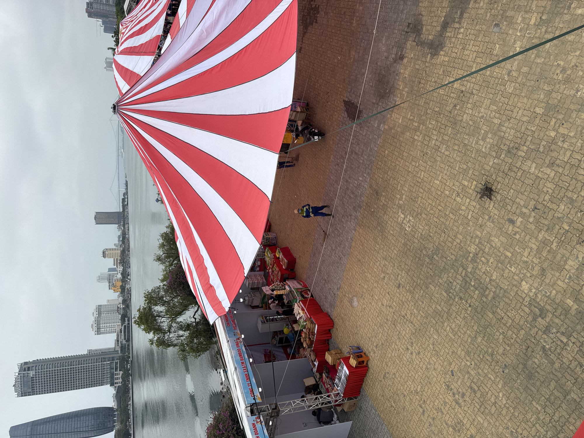 Here is a detailed description of the image:
The image captures a street market scene located in Đà Nẵng, Vietnam. The vantage point is from above, looking down at the market area which is covered by a large, striped red and white tent. In the background, the Hàn River and the skyline of Đà Nẵng are visible. A suspension bridge spans the river, connecting different parts of the city.
The market area consists of stalls, each displaying various goods on red-clothed tables. There are several people near the stalls; some appear to be vendors, while others may be customers. A person wearing a blue uniform and a face mask is walking across the paved area.
The foreground is composed of paving stones. A green rope stretches across the frame, possibly used to secure the tent. Overall, the scene depicts a bustling market atmosphere against the backdrop of Đà Nẵng's urban landscape.