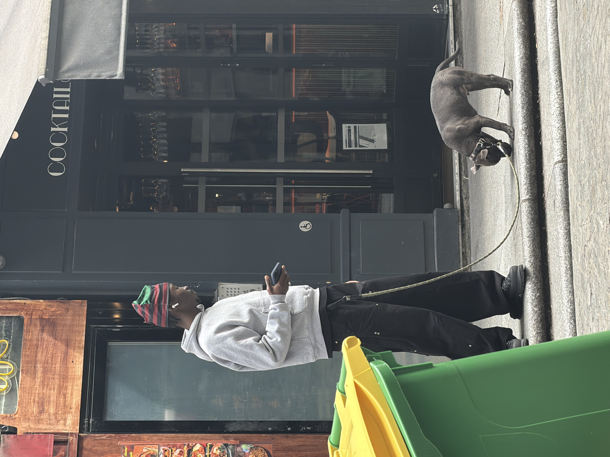 The image captures a scene in Paris, France. A young Black man is standing on the sidewalk looking at his phone. He is wearing a grey hoodie, black pants, and black shoes. He also has a colorful hat on his head and headphones in his ears. He holds the leash of a gray dog, possibly a pit bull, which stands on the sidewalk to his right. In the background is a building with a sign that says "COCKTAILS". Part of a green and yellow garbage container is visible in the foreground.