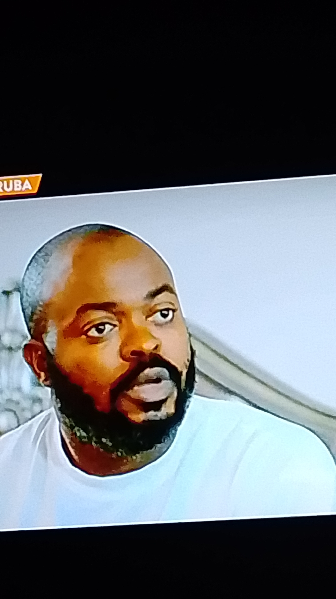 The image shows a close-up of a bald Black man with a beard, wearing a white t-shirt. He has dark eyes and appears to be looking off to the right. In the upper left corner of the image, a graphic element with the word "RUBA" in orange lettering on a black background is visible. The background behind the man is a light-colored wall and a blurred object that might be furniture. The man's skin tone is brown, and his beard is dark. His expression is neutral. The scene is indoors, with soft lighting. There is no specific indication of the city of Lokoja, Nigeria, in the visual content of the image.
