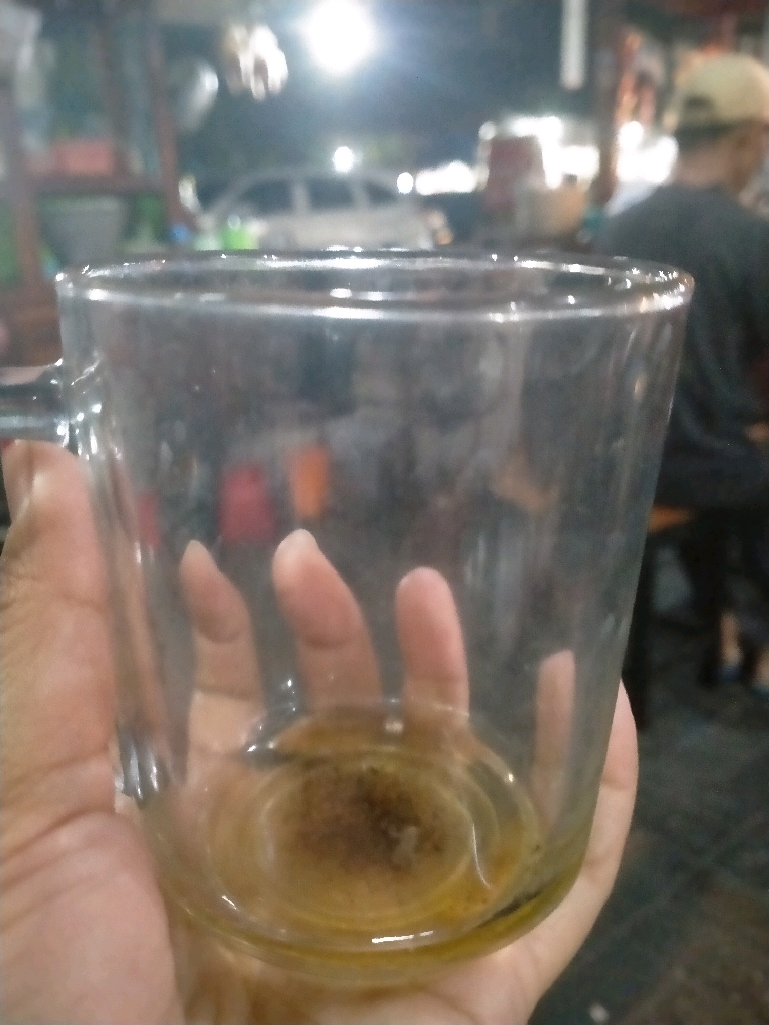 A close-up shot shows a person's hand holding a transparent glass mug. The mug contains a small amount of murky, yellowish liquid with sediment at the bottom. The background is blurred, suggesting an outdoor setting at night, possibly a roadside stall in Kasihan, Indonesia, given the presence of illuminated structures and what appears to be a parked vehicle. There are also out-of-focus figures of people in the background, one of whom is wearing a cap and dark clothing. The lighting in the scene is predominantly artificial, with bright light sources creating glare on the glass.