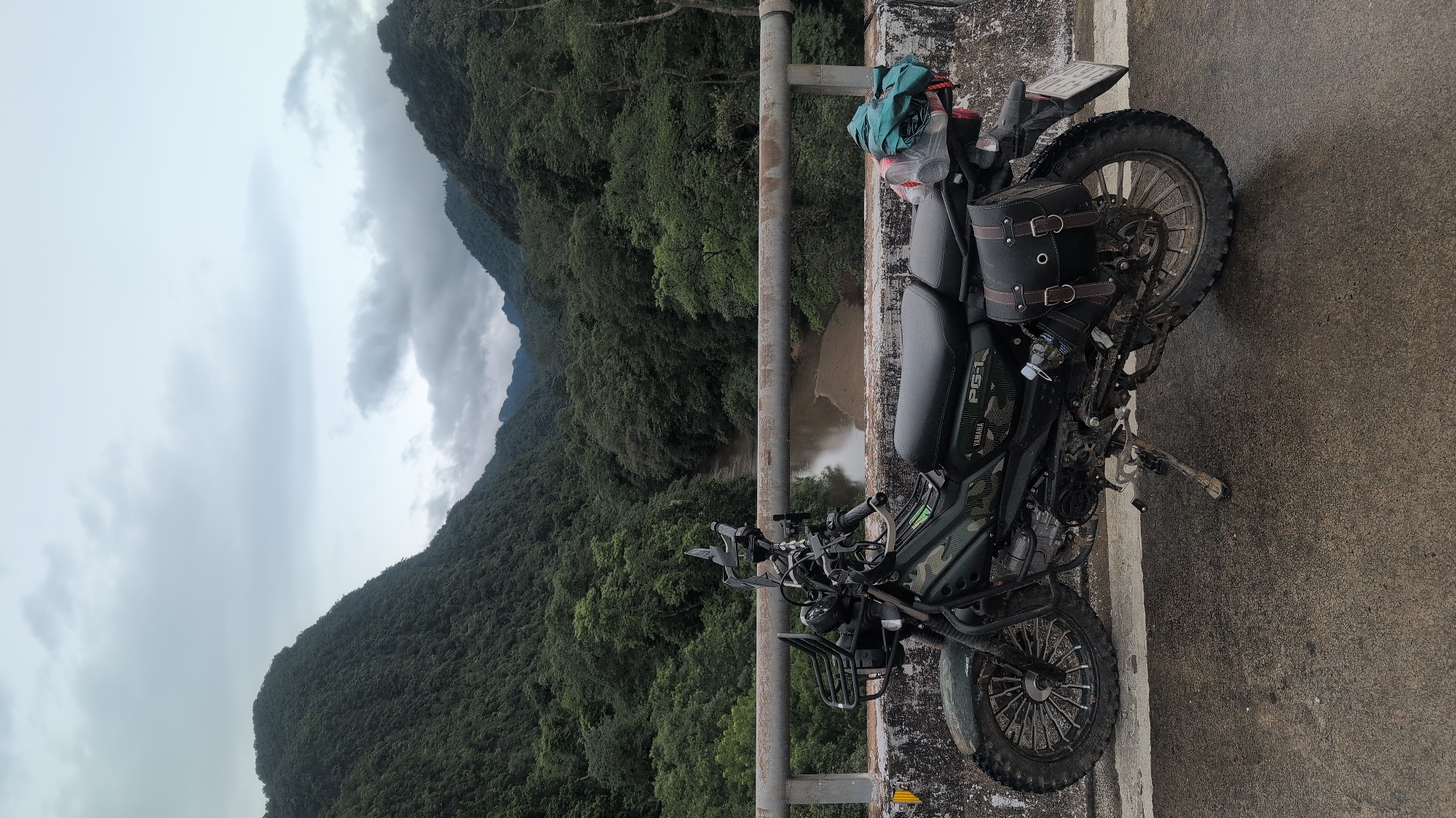 A motorcycle is parked on a bridge overlooking a river valley. The motorcycle is dark green with camouflage accents and has saddlebags and a basket on the front. It is facing to the right. The bridge has a concrete barrier and a metal railing. The valley is covered in lush green trees, and the sky is overcast. The location is Thượng Trạch Commune, Vietnam.