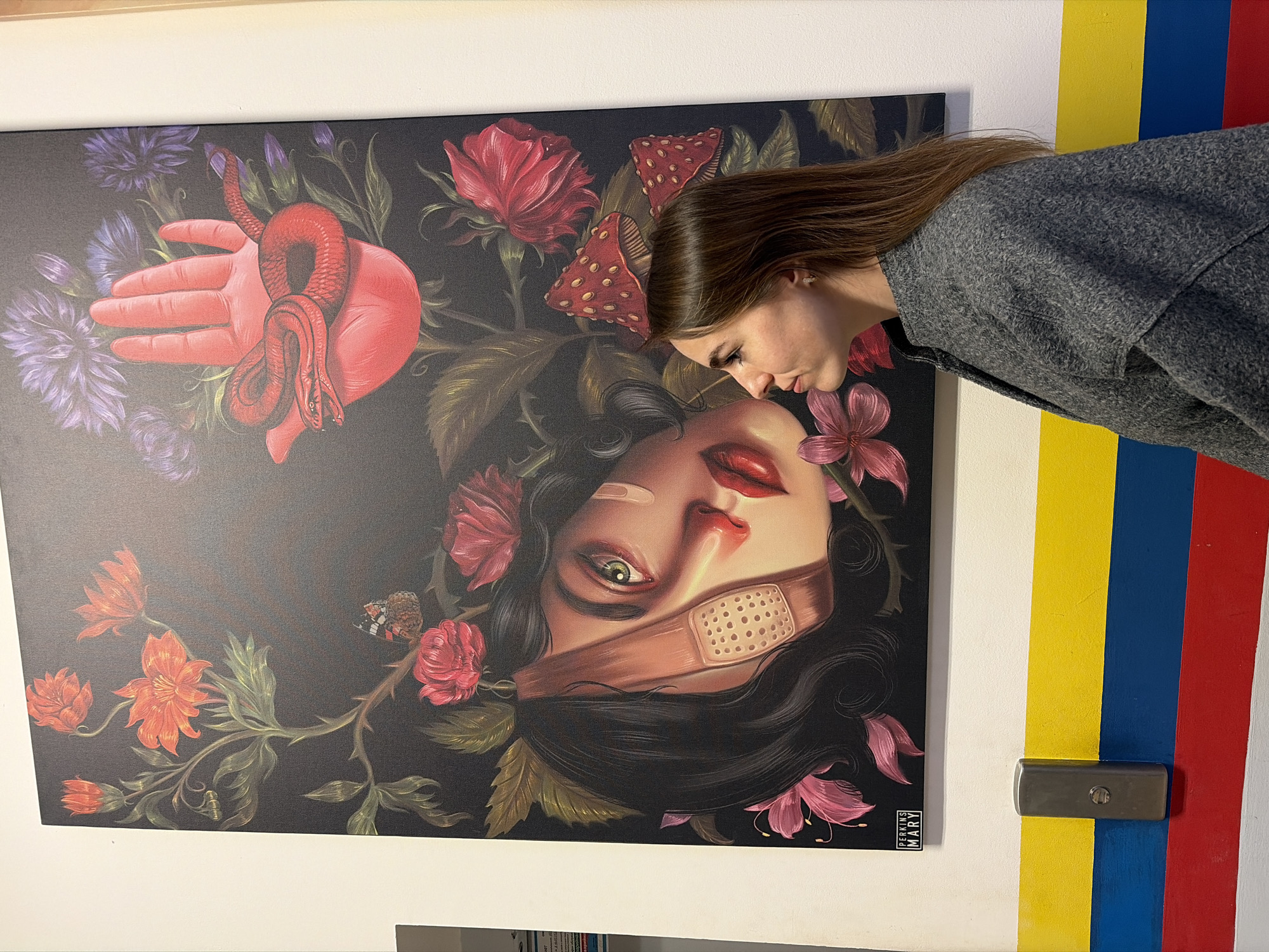 A woman with long brown hair stands next to a large, rectangular painting hanging on a wall. The painting depicts a woman's face partially covered by a band-aid, surrounded by flowers, a hand with a red snake, and mushrooms, all against a dark background. The wall features vertical stripes of white, yellow, blue, and red. A small silver box with a lock is also visible on the wall. The image was taken in the city of Izhevsk, Russia.