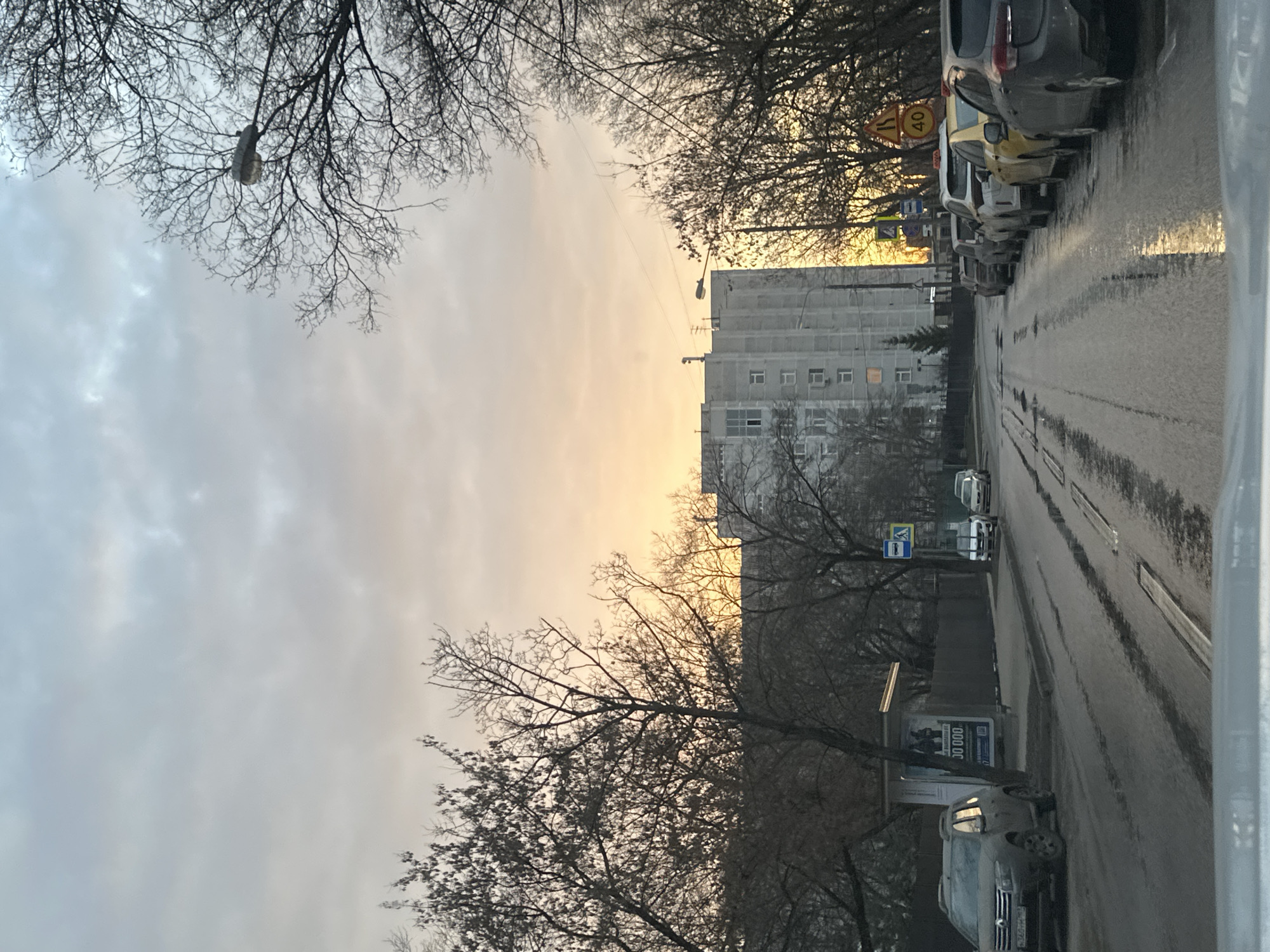 The image depicts a street scene in Moscow, Russia, on a cloudy day with a hint of yellow sunset visible in the distance. The street is wet, indicating recent rain or snow melt. Cars are parked along the right side of the road. A tall, grey apartment building is visible in the background, partially obscured by bare trees. Street signs are present, along with a bus stop or advertising structure on the lower right side of the frame.