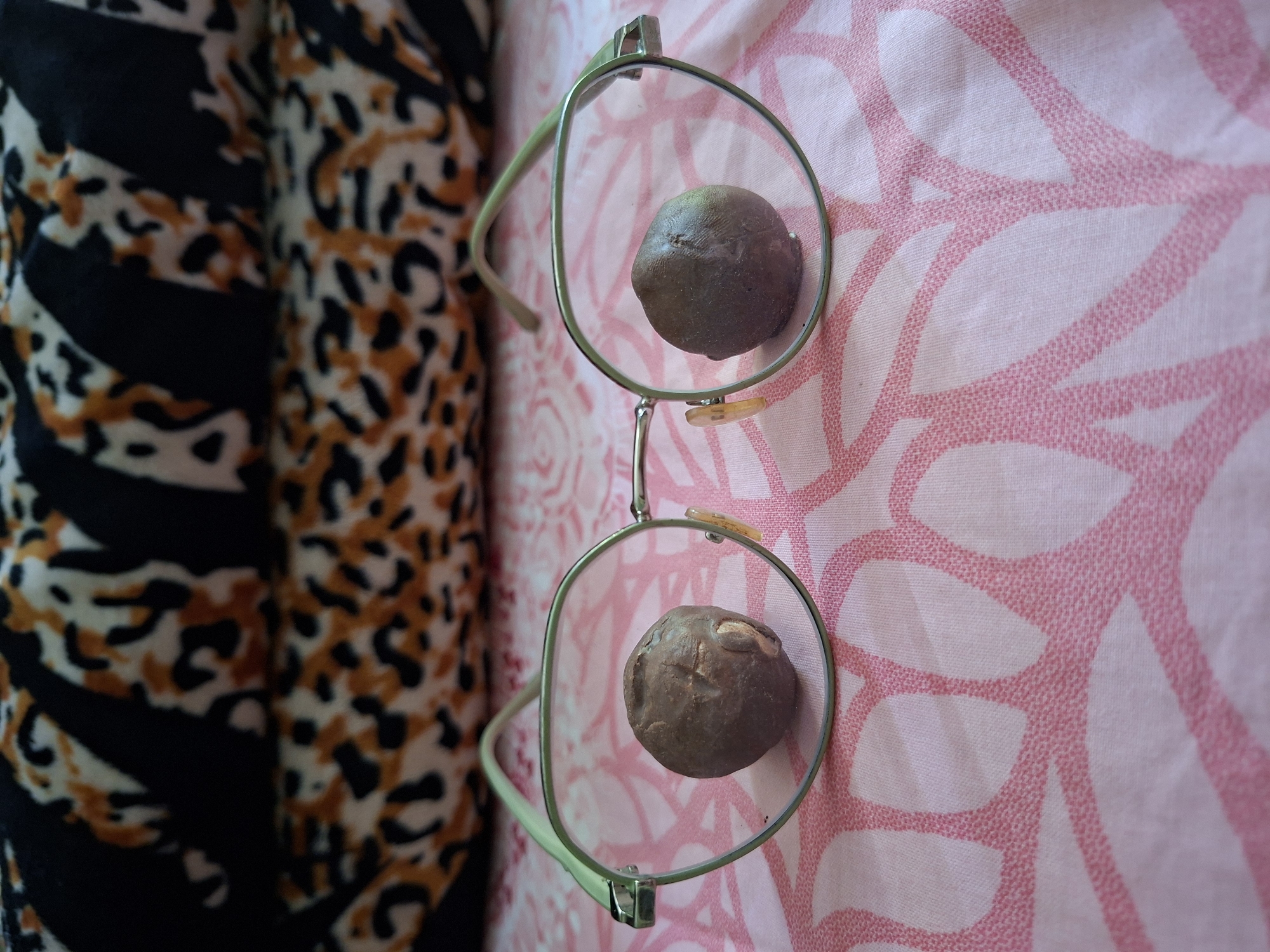 A pair of eyeglasses rests on a pink and white floral patterned fabric surface. The lenses of the glasses each contain a brown, spherical object, possibly stones. To the left of the glasses is a fabric with a black, white, and orange animal print. The location is specified as Kutaglumpang, Indonesia.