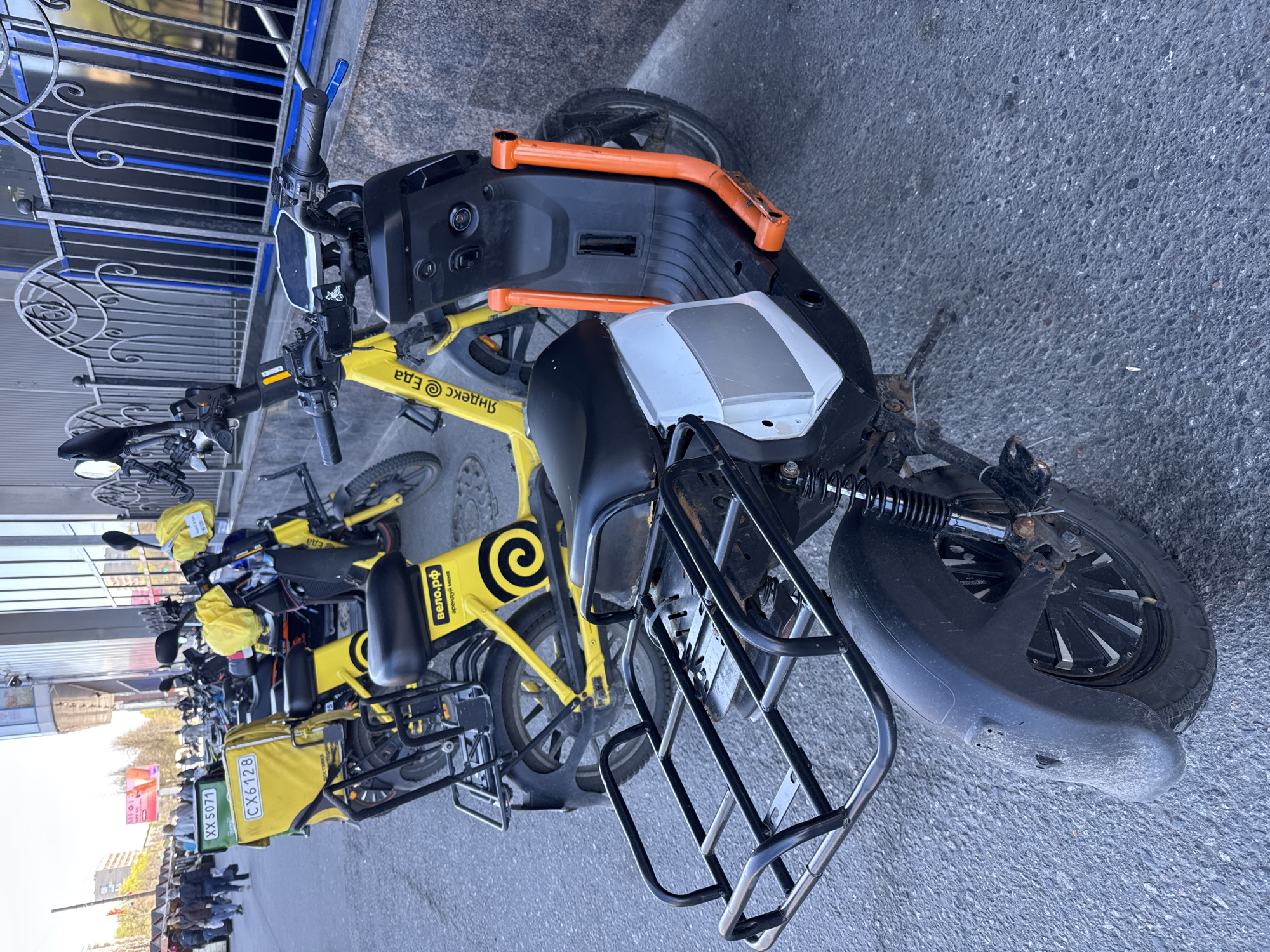The image shows a row of "Yandex.Eda" (Яндекс.Еда) electric bikes parked on the street. Yandex.Eda is a food delivery service. The bikes are yellow and black. Some have delivery boxes on the back. The license plate "XX5071" is visible on one of the delivery boxes. Some have yellow delivery bags with logos on them. In the background, there are people and buildings. The location is Saint Petersburg, Russia.