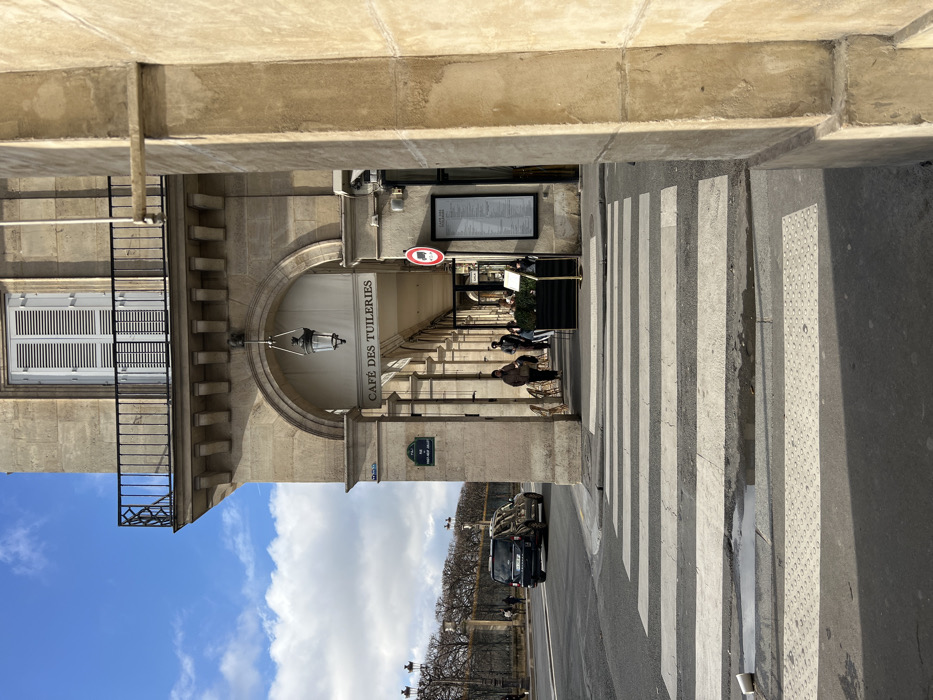 The image depicts a daytime street scene on Rue de Rivoli, Paris, France, adjacent to the Tuileries Garden. In the immediate foreground, a pedestrian crosswalk with alternating white and black stripes extends across an asphalt street. A white disposable cup rests on the crosswalk, next to a small puddle. To the right, a multi-story, light-colored stone building with classical architectural details features an arched arcade at ground level. A sign above the arcade identifies the establishment as "CAFÉ DES TUILERIES," and a black metal lantern is suspended from one arch. A blue street sign attached to a pillar reads "70 RUE DE RIVOLI." Under the arcade, approximately three individuals are visible: two figures walk away from the camera, and one or two patrons are seated at outdoor tables equipped with wicker chairs and a dark planter containing greenery. A red circular prohibitory sign with a white horizontal bar and a rectangular menu board are affixed to the cafe's facade. On the asphalt street, a dark-colored multi-purpose vehicle (MPV) is driving away from the viewer. The background reveals bare deciduous trees and ornate streetlights, indicative of the Tuileries Garden. The building's upper level includes a balcony with a black wrought-iron railing, positioned below a window fitted with white louvered shutters. The sky overhead is partially cloudy.