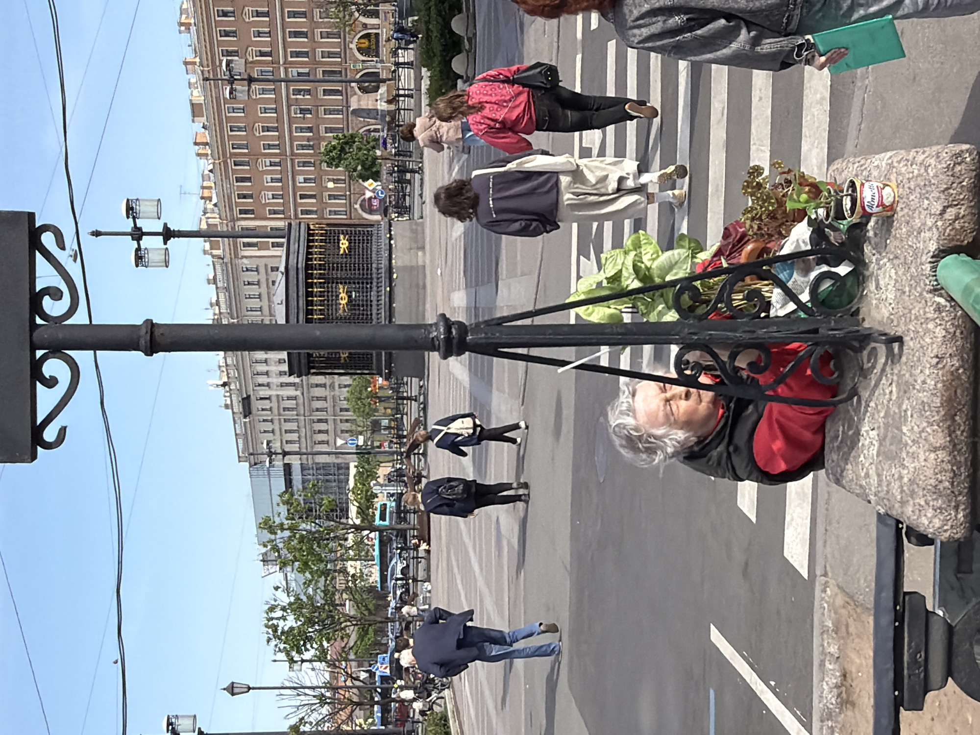 The image depicts a street scene in Saint Petersburg, Russia. Pedestrians are crossing a crosswalk in front of a large building with numerous windows. A woman in a red shirt and dark jacket is standing next to a stone structure with plants in metal cans. There is an ornate lamppost on the left side of the image. Other pedestrians are walking in different directions. The sky is blue and clear.