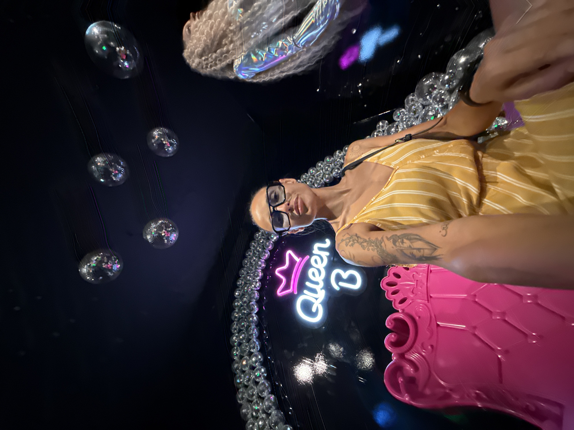A young woman with dark hair and large black-rimmed glasses is taking a selfie in a dimly lit, vibrant space. She is wearing a yellow and white striped sleeveless dress and has tattoos on her arm. She is holding her phone to take the picture, with her right arm extended.
To her left, a bright pink, ornate chair resembling a throne is visible. Behind her, a neon sign glows, spelling out "Queen B" with a crown above the word "Queen." The background is adorned with numerous reflective, spherical decorations, appearing like disco balls, some of which are hanging from the ceiling and others forming arches or structures around the scene.
To the right of the woman, a mannequin or display with long, curly blonde hair and a shimmering, iridescent silver outfit is partially visible. The overall ambiance suggests an entertainment venue or a themed photo opportunity, possibly a club or a themed bar. The lighting is dramatic, with spotlights and the neon sign creating a lively and somewhat theatrical atmosphere.
The image appears to be taken in Prague, Czechia, given the context of the request.
