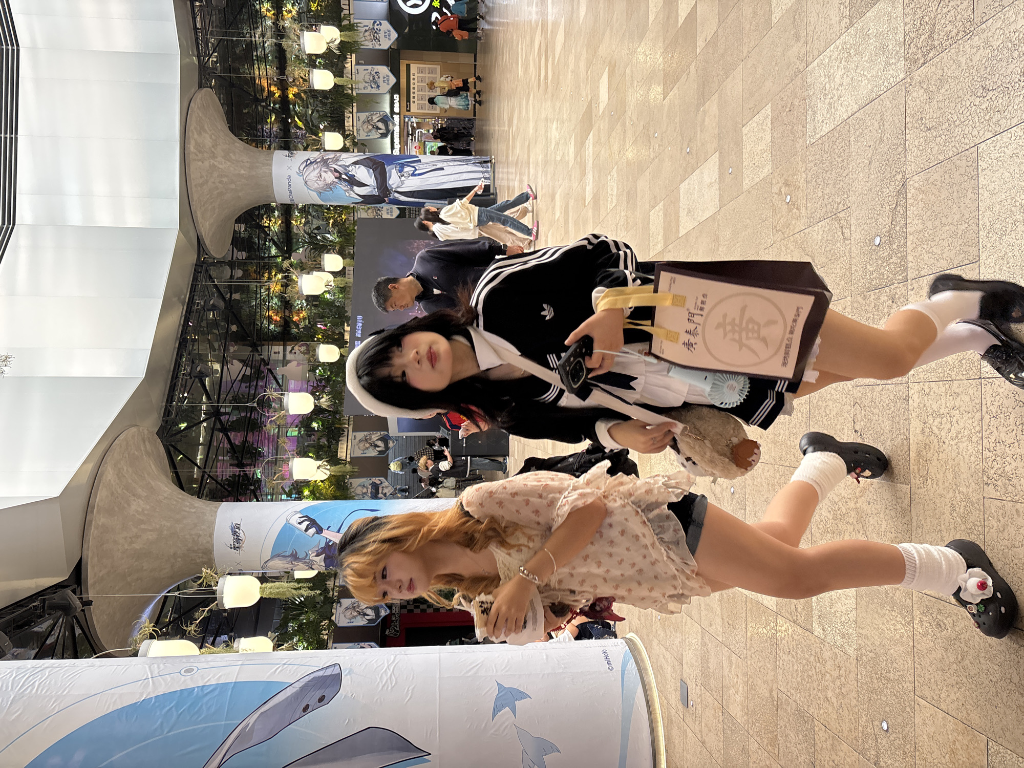 Two young women stand inside a building in the Liangjiang New Area of Chongqing, China. One woman, wearing a black Adidas track jacket over a white shirt, a white headband, and black shoes, is holding a tan stuffed animal and a paper bag with Chinese writing. The other woman, with blonde hair, wears a floral-patterned shirt, black shorts, and black shoes with white socks. They are standing in front of large cylindrical displays with anime characters printed on them. The background includes indoor plants, light fixtures, and other people. The floor is tiled.