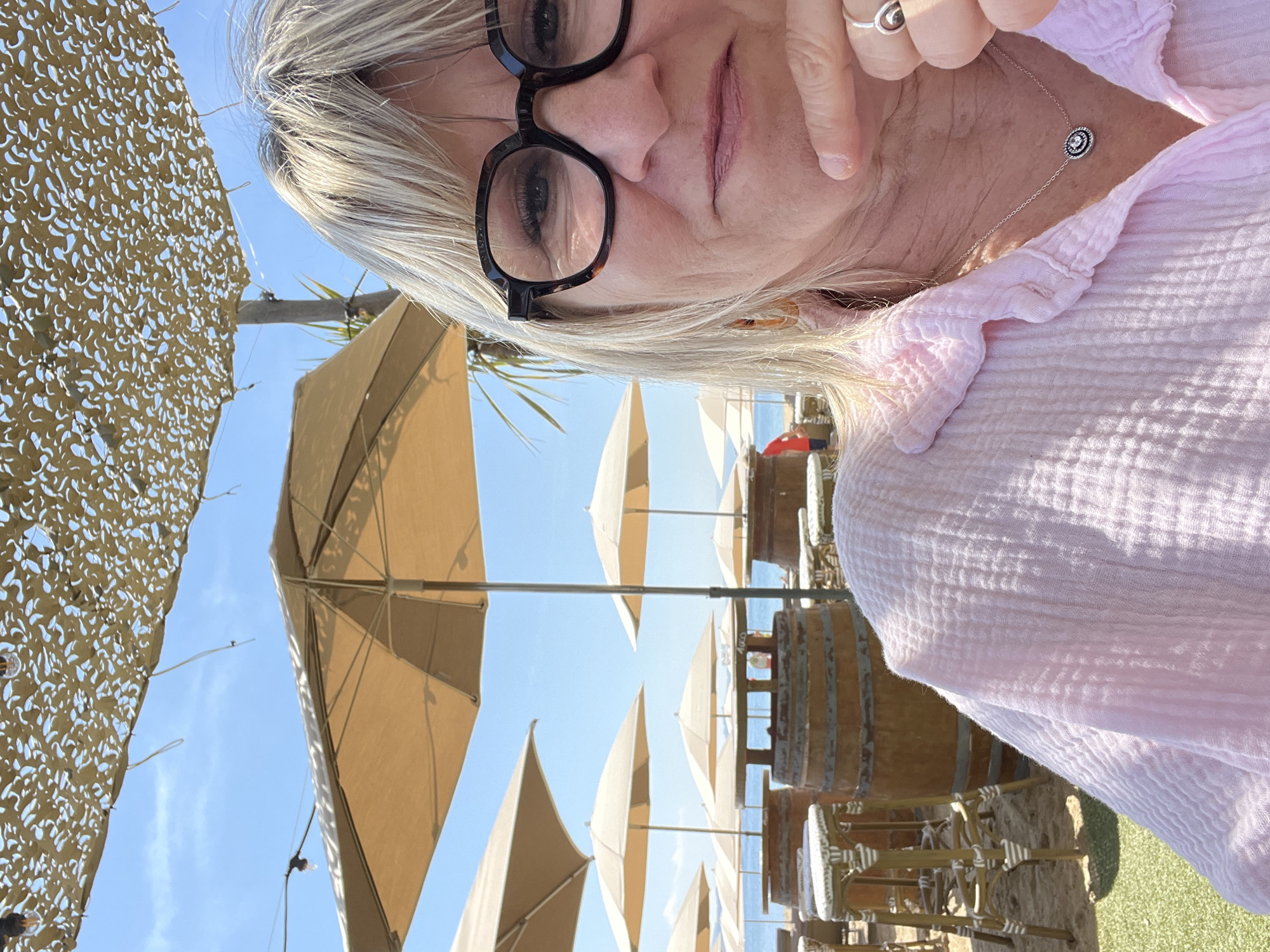 A woman wearing glasses and a light pink shirt takes a selfie outdoors at a café or restaurant in Sainte-Maxime, France. The background features multiple beige sun umbrellas, wooden tables made from barrels with chairs, and a blue sky. Camouflage netting is visible on the left. The woman is wearing a ring and a necklace. The sea is visible in the distance.