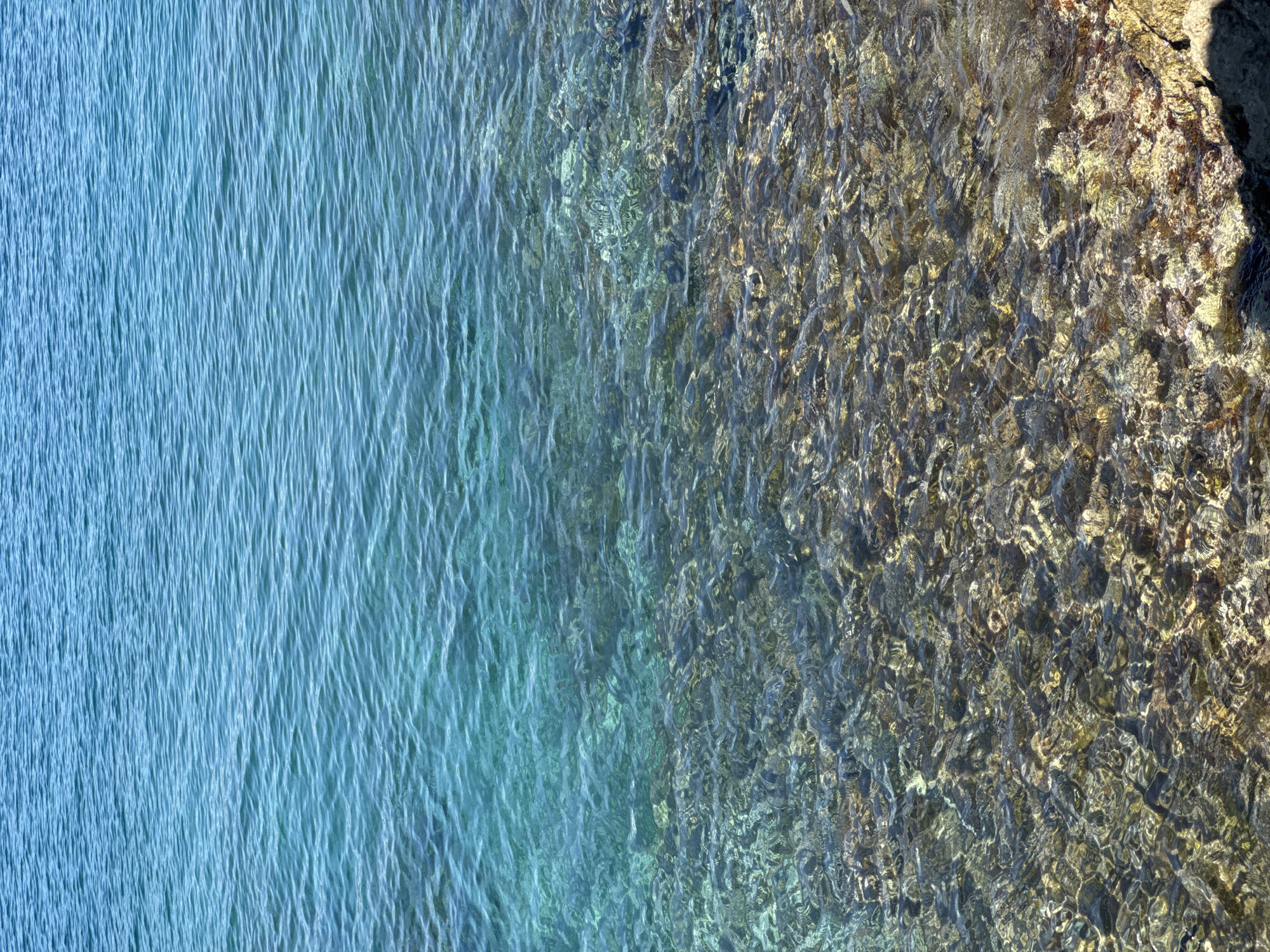 The image shows a close-up view of shallow, clear turquoise water over a rocky seabed. The water's surface is rippled, catching the sunlight and creating a shimmering effect. The seabed is composed of numerous submerged rocks of varying sizes and colors, predominantly grey, brown, and yellowish-tan, with some appearing smoother and others more irregular. The transition from deeper, more uniformly blue water at the top of the frame to the shallower, clearer water revealing the seabed occurs roughly in the middle of the image. The bottom right corner shows a sliver of the shore, with larger, sunlit rocks. The scene is devoid of any people or man-made objects, focusing solely on the natural underwater environment. This location is the sea in Dubraljevina, Montenegro.