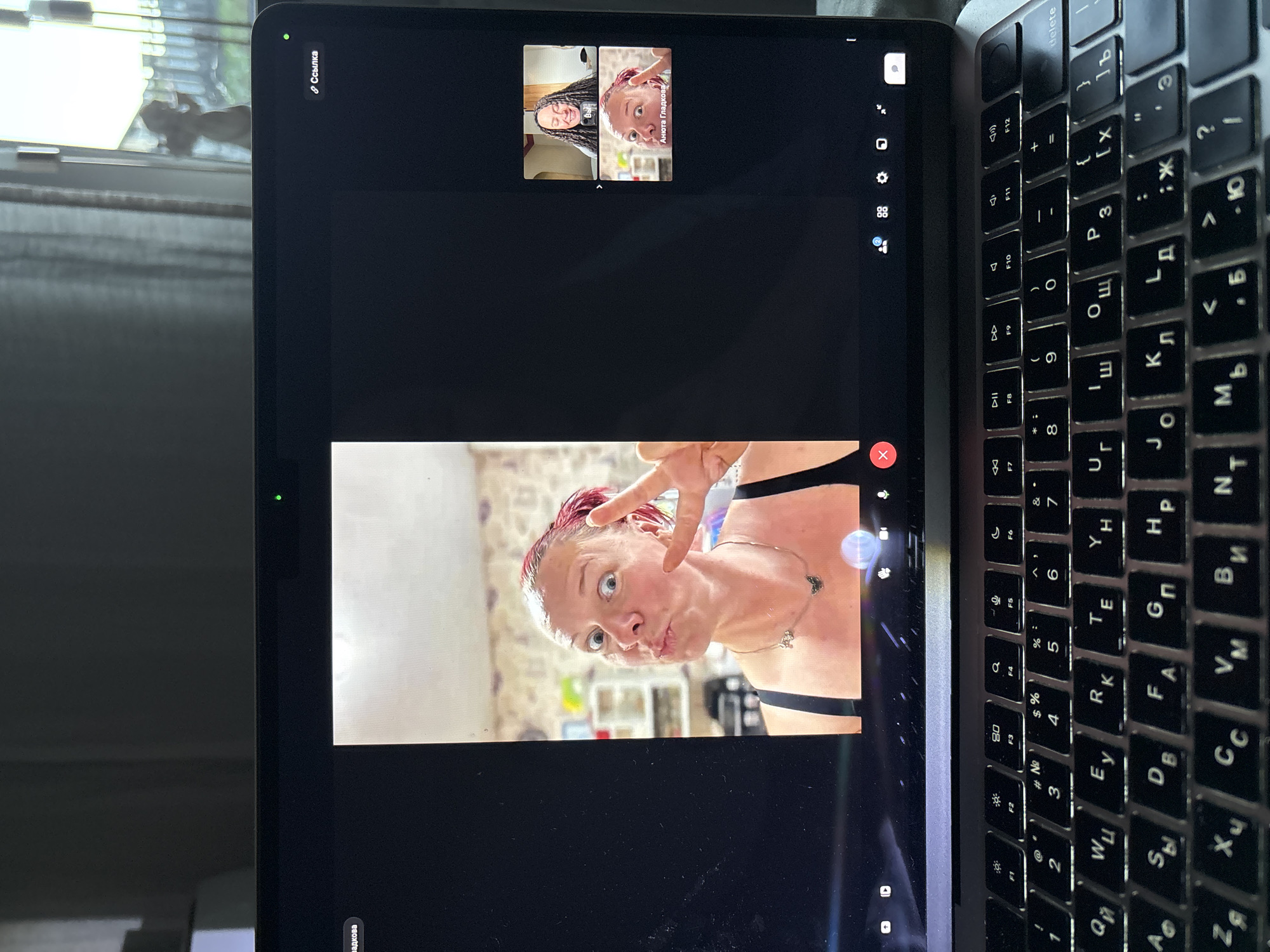 An image captures a laptop displaying a video call in a slightly rotated, vertical orientation. The main screen shows a woman with reddish hair making a playful face, pouting her lips, and flashing a peace sign with her fingers. She is wearing a dark top, and a light-colored room with shelves is visible in her background.
In smaller windows at the top of the screen, two other participants are visible. One, labeled "ВЫ" (You), shows a woman with dark, curly hair smiling. The other, labeled "Анюта Гладкова" (Anyuta Gladkova), shows a person with a similar appearance to the main screen participant. A green light is illuminated at the top left edge of the laptop screen, indicating the camera is active.
The laptop's keyboard, featuring both Latin and Cyrillic characters, is visible on the right side of the image, suggesting a Russian-speaking user. Keys such as "Delete," function keys F1-F12, and various alphanumeric keys in both layouts are discernible. The surrounding environment appears to be indoors, with natural light coming from a window with blinds visible behind the laptop.
The visible text on the screen includes "Ссылка" (Link) at the top left, "ВЫ" (You) on one small video feed, and "Анюта Гладкова" on another. The location context provided is Chaikovsky, Russia.