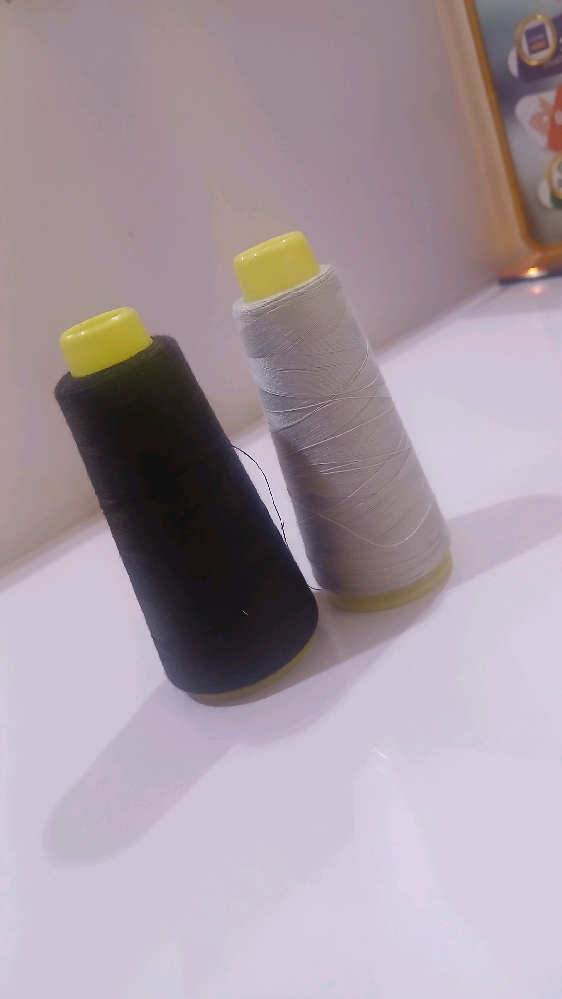 Two spools of thread are shown, one black and one light gray, against a neutral background. The spools have yellow caps. The black spool is in the foreground and is tilted to the left. The gray spool is behind and to the right of the black spool, also tilted. A thin black thread extends from the black spool. A framed object, possibly a picture or decorative item with cartoon characters, is visible in the upper right corner, out of focus. The surface beneath the spools appears to be white and smooth, reflecting some light. There is a shadow cast by the black spool to its lower left. The scene is indoors, with soft, diffused lighting. There is no information available regarding the location in the city of Gura Top, Nigeria.