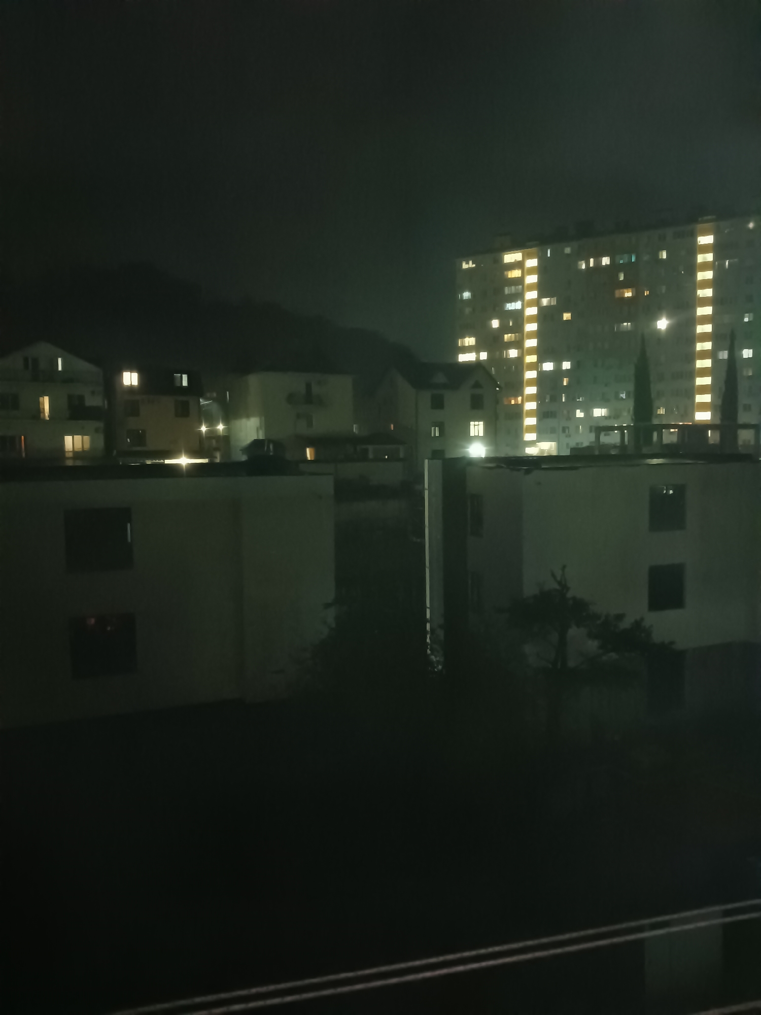 The image captures a nighttime view of a city, likely Sochi, Russia, with several buildings illuminated by interior lights. In the foreground, two lower, light-colored buildings with dark rectangular windows are prominent. Behind these, a cluster of smaller residential buildings with illuminated windows is visible, set against a dark, hazy sky that partially obscures a mountainous backdrop. To the right, a tall, multi-story building with a grid of glowing windows stands out, its bright lights contrasting with the surrounding darkness. Several slender, dark trees, possibly cypress or fir, rise between the buildings. The overall atmosphere is dark and slightly ominous, with the artificial lights piercing the night.