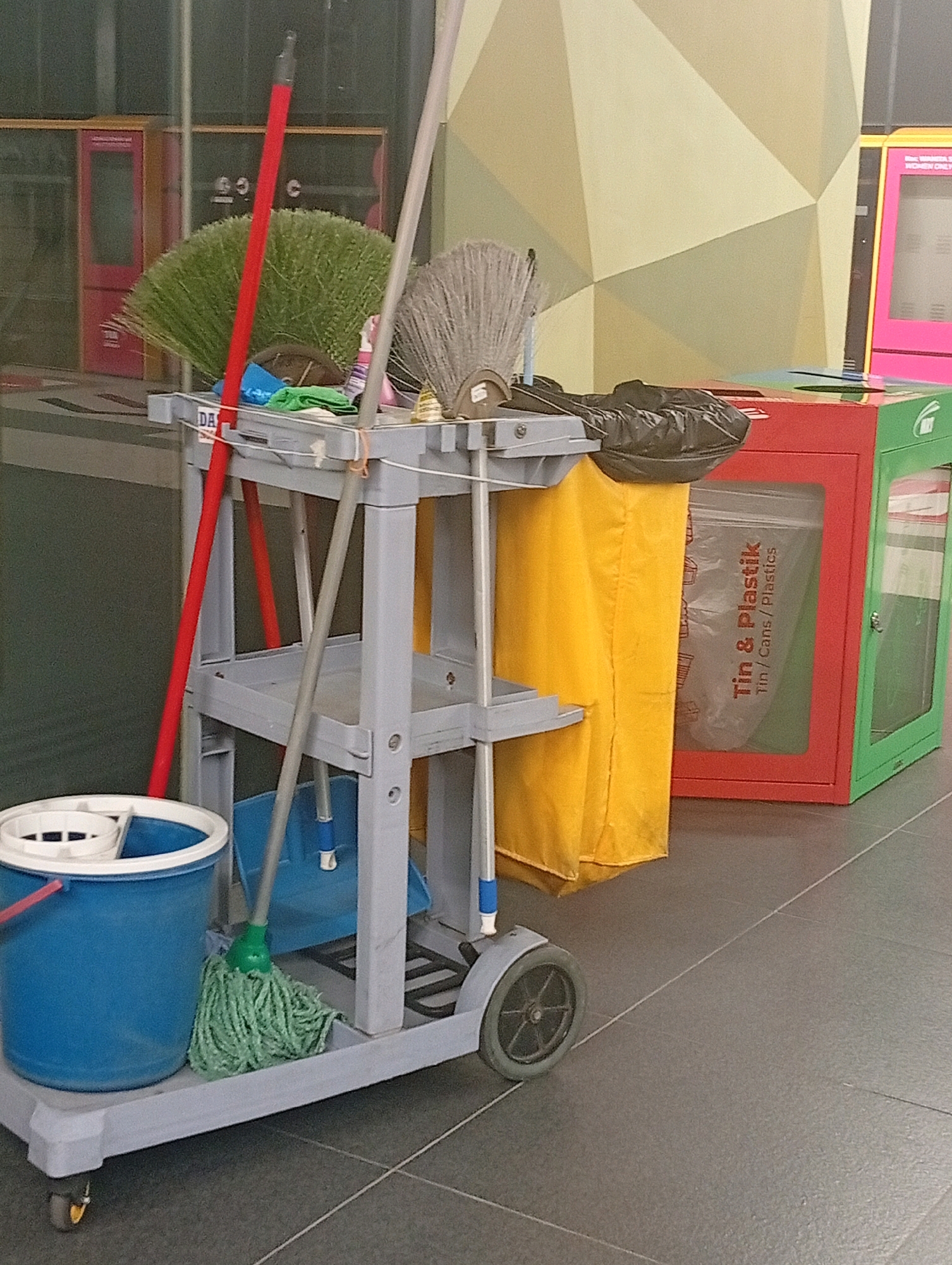 An indoor scene shows a cleaning cart with various cleaning supplies and waste receptacles. The cart, made of light gray plastic, is equipped with a blue bucket, a green mop, a dustpan, brooms, and a yellow laundry bag. Behind the cart, a red and green waste bin with the label "Tin & Plastik / Tin/Cans/Plastics" is visible. The background features a wall with geometric patterns and two pink doors. The floor is tiled in a dark gray color. This scene is located in the basement of the shopping mall complex of Paradigm Mall in Petaling Jaya, Malaysia. There are no people visible in the image.