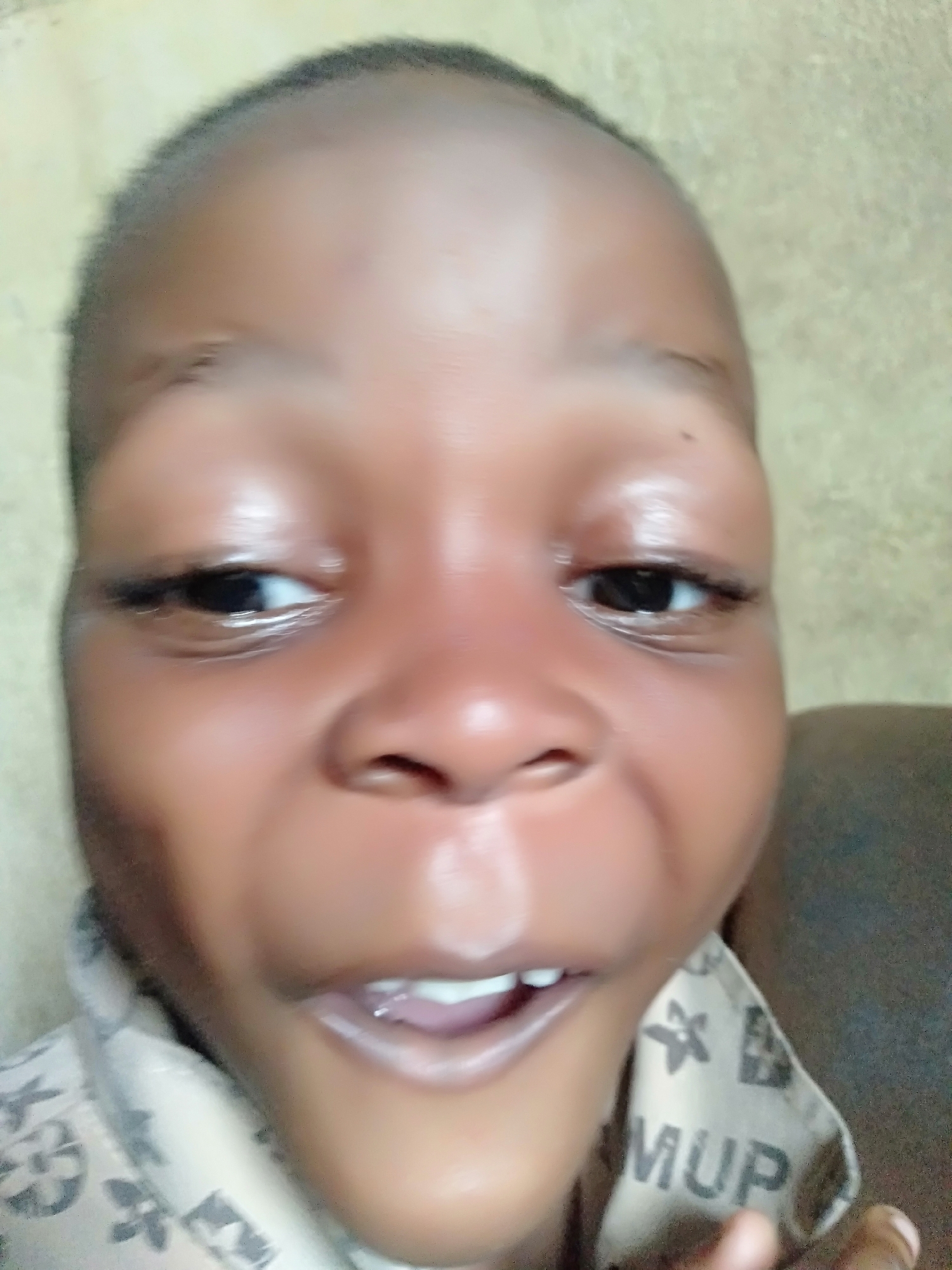 A close-up shot focuses on the face of a child with dark skin and short hair, wearing a patterned shirt. The background is out of focus, but appears to be an interior space. The location is unknown.