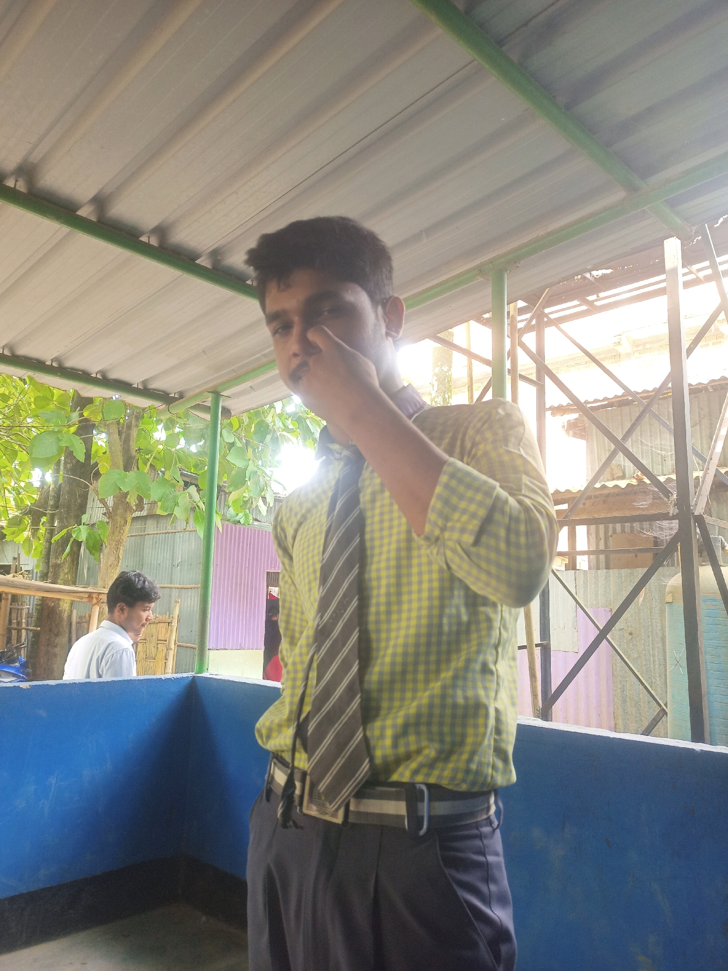 The image depicts a young male, likely a student, standing in an outdoor setting under a corrugated metal roof supported by green metal poles. He is wearing a yellow and grey checkered shirt, a striped tie, and dark trousers with a striped belt. His right hand is raised to his nose, and he is looking directly at the camera. In the background, another young male in a white shirt is visible, looking to his left. Behind them, there are trees with green leaves, a corrugated metal wall, and metal scaffolding. The ground appears to be concrete. The lighting suggests it is daytime, with sunlight filtering through the roof and the scaffolding. The presence of a school uniform on the main subject and the overall environment suggest a school or community area in Hatsinigmari, India.