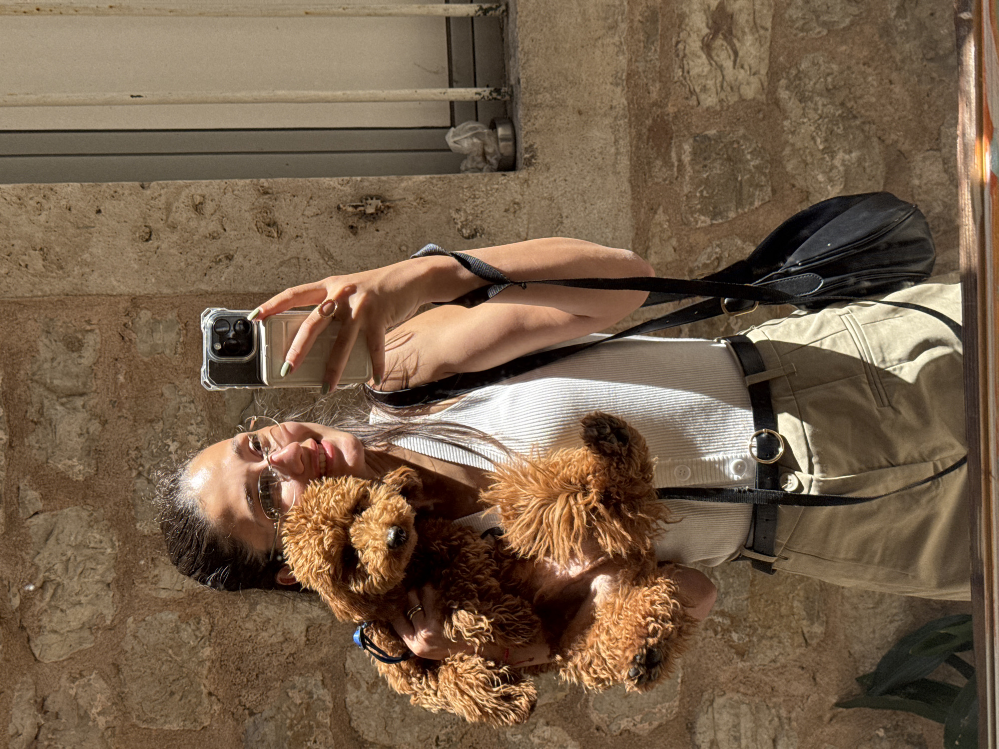 A young female, approximately 20-30 years old, is depicted taking a selfie outdoors in Budva, Montenegro. She is smiling, wearing clear-rimmed glasses, a white ribbed tank top, khaki trousers, and a black belt with a gold circular buckle. Her fingernails are painted light green, and she wears a gold ring on her right hand holding a smartphone in a clear case, and another gold ring on her left hand. She holds a small, curly-haired, orange-brown poodle-type dog, wearing a blue harness, in her left arm. The dog is facing slightly to the right, towards the camera. The background consists of an old stone wall constructed from irregularly shaped, light brown stones with visible mortar lines. To the upper right, a window with three vertical white metal bars is partially visible, below which a small metal bowl contains crumpled white material. Direct sunlight illuminates the scene from the left, creating distinct highlights and shadows on the subjects and the stone wall. A dark wooden ledge or railing is partially visible at the bottom of the frame, with a subtle multicolored reflection.