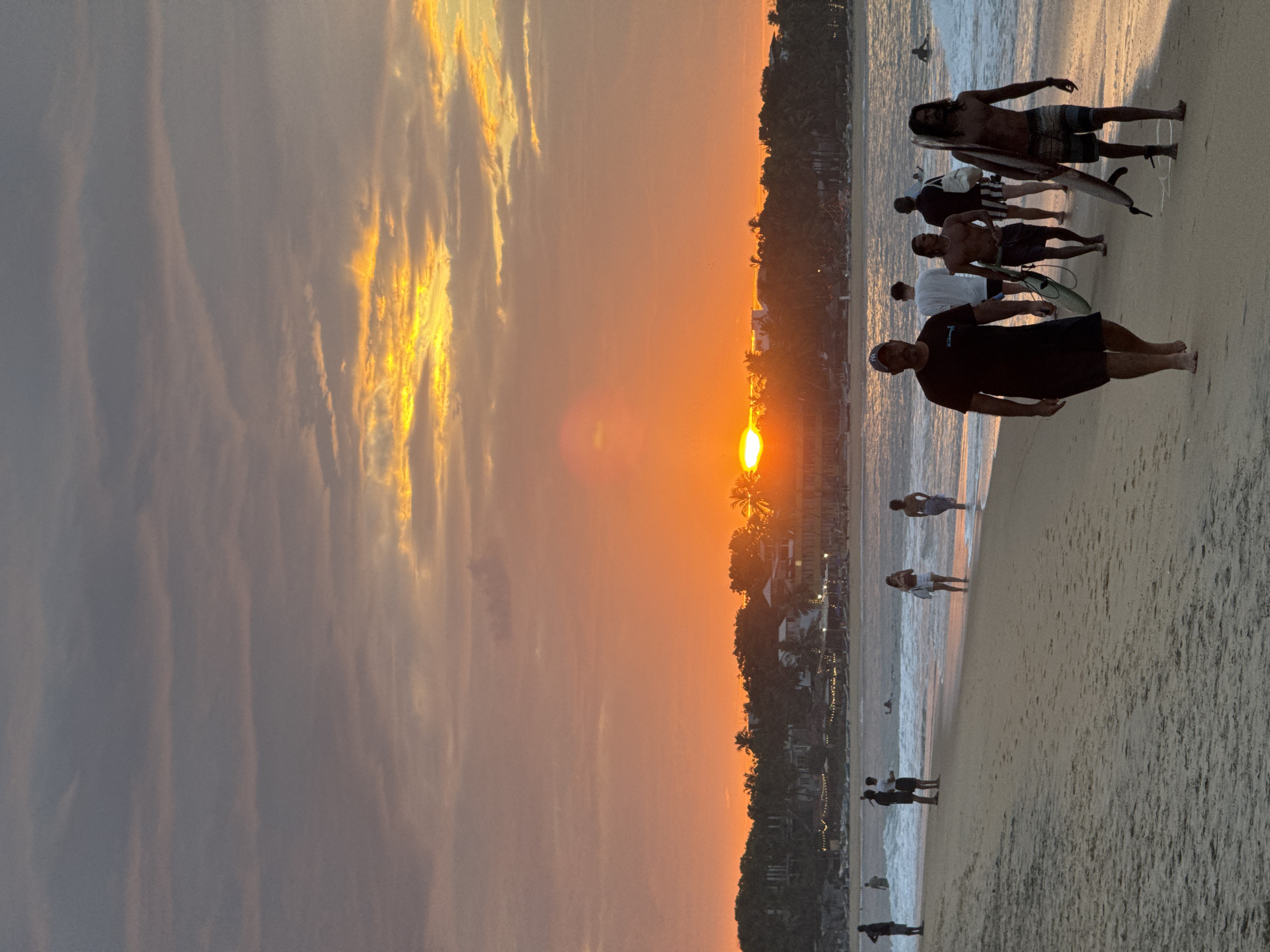 The media file captures a sunset scene on a beach. Several people are present on the sandy shore and at the edge of the water, some with surfboards. The setting is a coastal area with buildings and palm trees visible in the background, suggesting a tropical location. The sky is dominated by warm, vibrant colors of orange and yellow, characteristic of a sunset. Wispy clouds are scattered across the sky, some illuminated by the setting sun. The mood appears relaxed and recreational, with individuals seemingly enjoying the end of the day at the beach. The time of day is clearly sunset, given the lighting and sky colors. The location cues point towards a beach in Arugam Bay, Sri Lanka. There is no visible text in the media file.