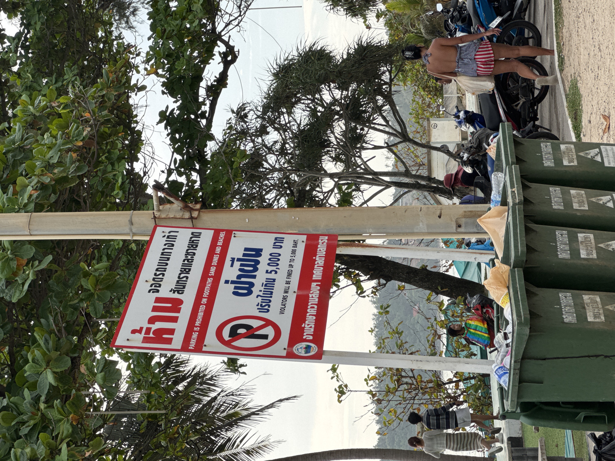 A close-up shot captures a signpost in Karon, Thailand, displaying multilingual warnings about parking. The sign, in red and white, bears Thai script and English text stating: "PARKING IS PROHIBITED ON FOOTPATHS SAND DUNES AND BEACHES. VIOLATORS WILL BE FINED UP TO 5,000 BAHT." Below this, a circular "P" symbol with a diagonal line across it is prominently featured, indicating "no parking." A seal with the text "จวนรักษาความสงบ เทศบาลตำบลกะรน" (roughly translates to "Peacekeeping Department, Karon Subdistrict Municipality") is visible on the lower part of the sign. In the foreground, two large green trash bins are filled with various items, including plastic bottles and bags, with one bin showing a recycling symbol. Behind the bins, several motorcycles are parked, and a woman in a checkered top and striped shorts walks away from the camera. Further in the background, two individuals are walking away, and others are seated, with a body of water visible beyond them. The scene is set against a backdrop of lush green trees and a cloudy sky, suggesting an outdoor, possibly beachside, location in Karon, Thailand.