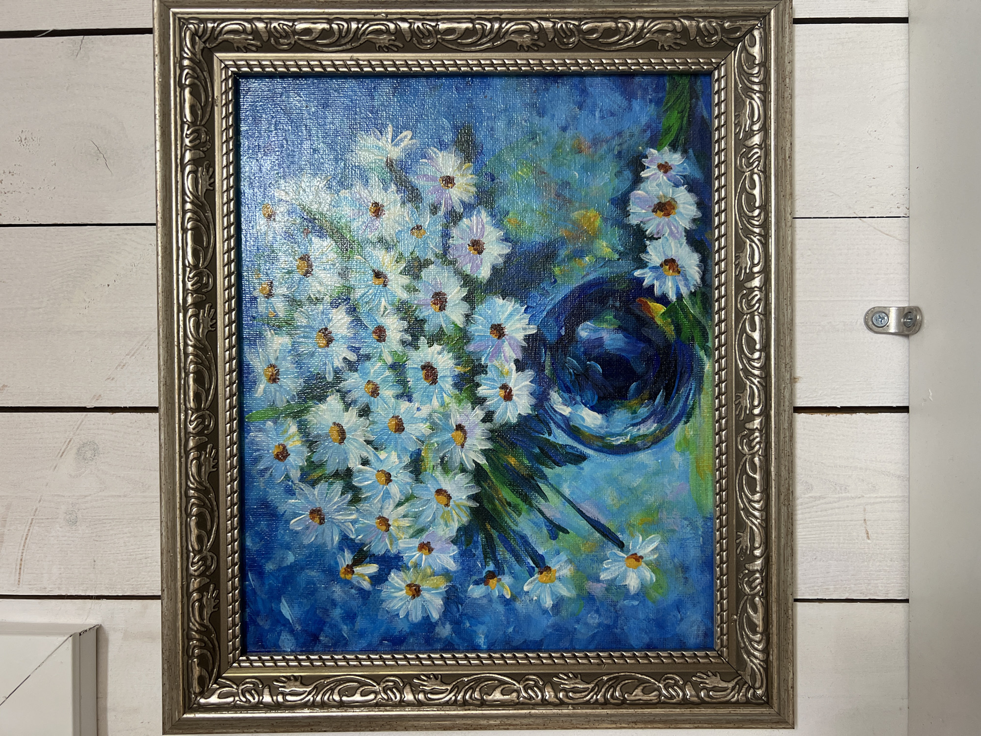 This is a photograph of a framed oil painting of daisies in a vase, hung against a white wooden wall. The painting features a bouquet of white daisies with yellow centers against a vibrant blue background. The flowers are rendered with visible brushstrokes, giving them a textured appearance. A dark blue, reflective vase holds the bouquet. The frame is ornate and appears to be silver or pewter in color. The white wooden wall behind the painting is composed of vertical planks. There is no indication of the city of Angarskie Khutora, Russia, in the image.