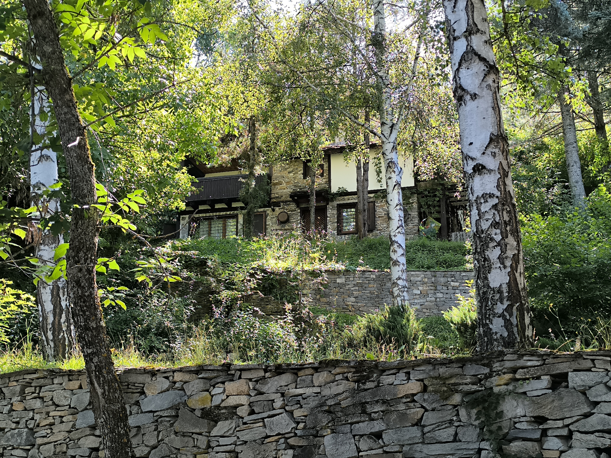 The image shows a stone-built house nestled amongst lush green trees, likely in the ethno-architectural village of Kovachevitsa, Bulgaria. The house features a traditional architectural style with stone walls and wooden accents, partially obscured by the dense foliage. A dry-stone wall forms a barrier in the foreground, showcasing the rugged beauty of the natural surroundings. Sunlight filters through the leaves, casting dappled shadows and highlighting the textures of the stone and bark. The scene conveys a sense of tranquility and immersion in nature, characteristic of rural Bulgarian villages.