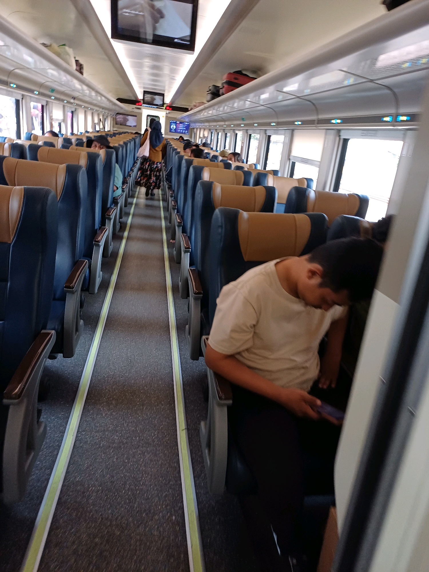 The image shows the interior of a train carriage with rows of empty and occupied seats on either side of a central aisle. The seats are upholstered in blue and tan. The flooring is a dark grey carpet with yellow and white markings along the edges of the aisle. Overhead compartments run along the length of the carriage.
In the foreground, a young person wearing a light-colored t-shirt and dark pants is seated and leaning their head against the seatback, appearing to be asleep or resting. They are holding a smartphone in their right hand.
Further down the aisle, a woman wearing a hijab and a patterned skirt is walking away from the camera. Several other passengers are visible in the seats, some of whom are looking at their phones or resting.
Two digital screens are mounted on the ceiling of the carriage, displaying visual information. Luggage, including a red suitcase, is stored in the overhead compartments. The lighting in the carriage appears to be a combination of overhead fluorescent lights and ambient light from the windows.
The image was captured on a train traveling within or near Surakarta, Indonesia.
