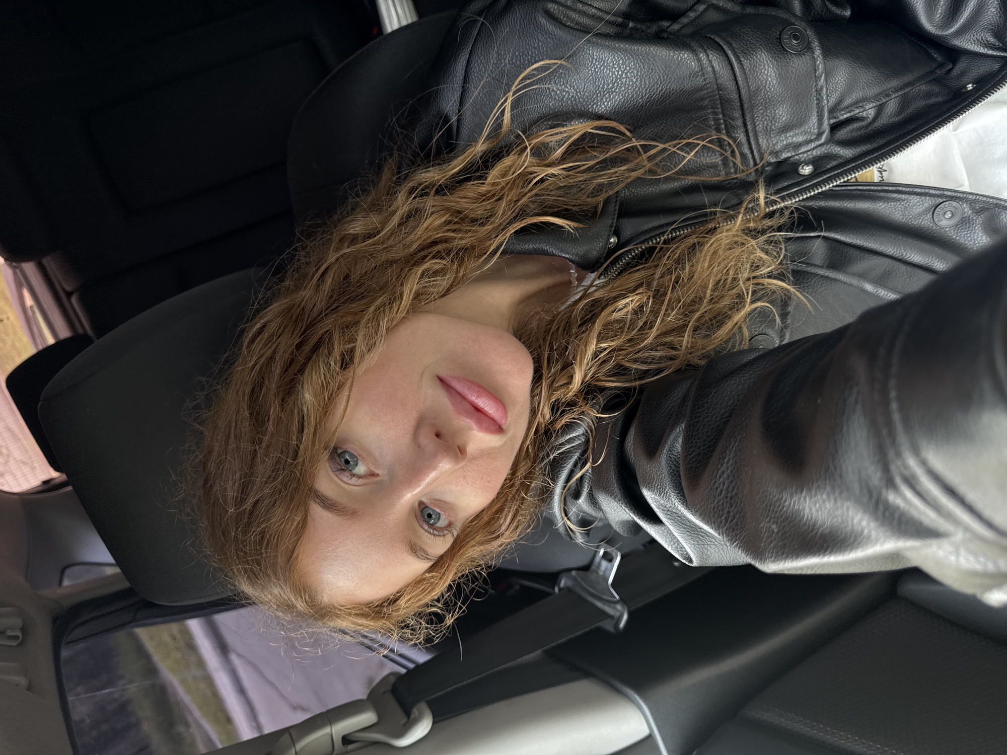 A female individual is depicted in a close-up selfie taken inside the front passenger seat of a vehicle. She has long, wavy, reddish-blonde hair and blue eyes, looking directly at the camera. Her skin tone is fair, and she has light pink lip color. She is wearing a black faux-leather jacket with a zippered front, metal snaps on the collar, and flap pockets on the chest, over a white inner garment and a faint, light-colored necklace. A dark fabric seatbelt is visible across her chest. The car's interior is dark, with a dark headrest behind her and dark upholstered rear seats visible in the background. Through the car window, a blurred outdoor environment is visible, featuring a paved surface, a distinct grey curb or barrier, and green foliage, indicating natural daylight conditions. There are no discernible features in the image to identify the specific location as Chaikovsky, Russia.