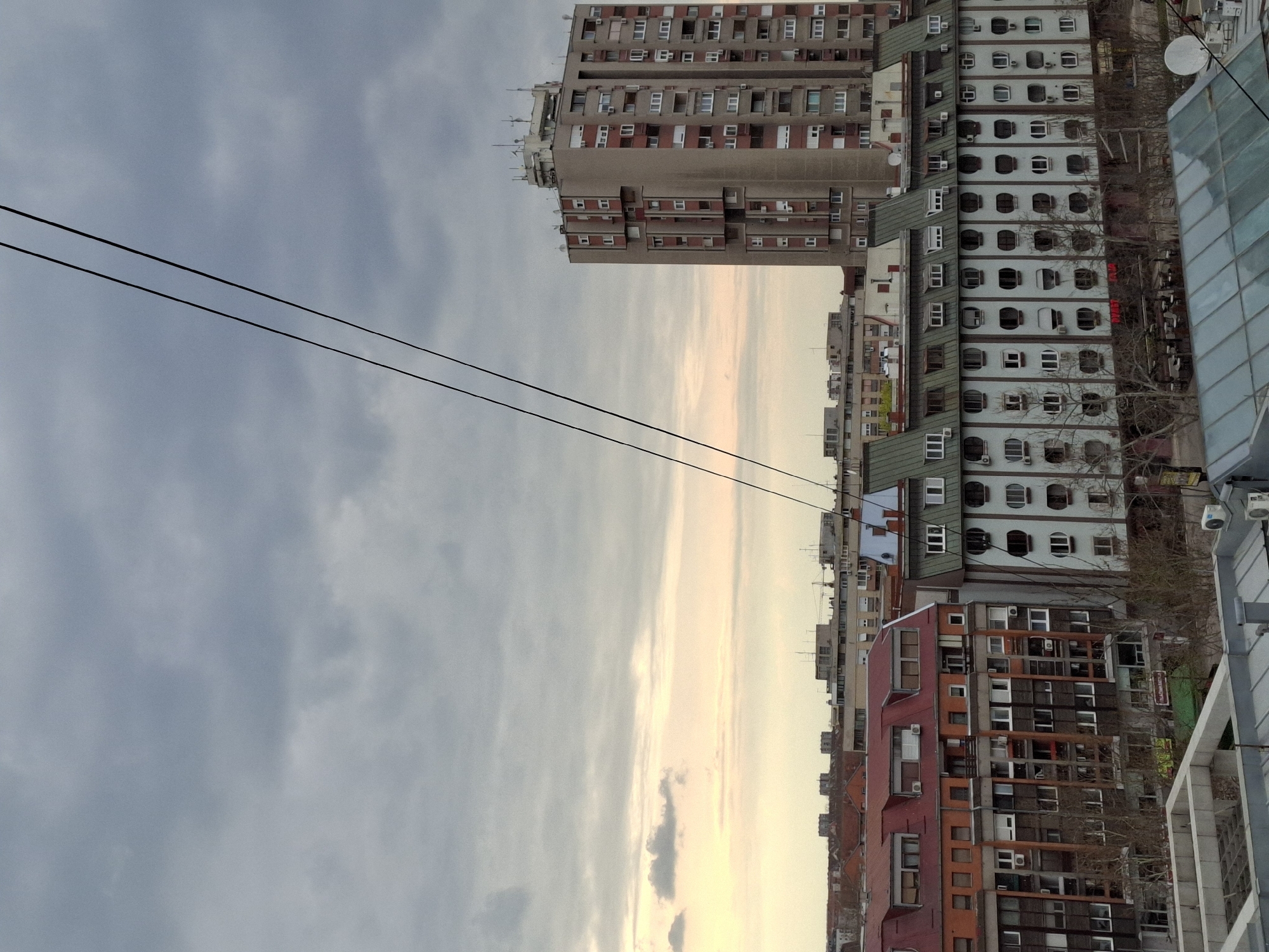 The photograph depicts a cityscape scene in Novi Sad, Serbia. The focal point is a cluster of residential buildings of varying heights and architectural styles. A large, gray-beige apartment complex, topped with communication equipment, stands out prominently. Adjacent to it is a light-blue building with distinctive arched windows and green roofs. A red and brown building with a steeply-sloped roof occupies a lower section. The sky is filled with striated clouds and hints of a light-yellow sunset, suggesting the photo was taken during the late afternoon or early evening. Electrical wires are suspended in the upper-left quadrant.