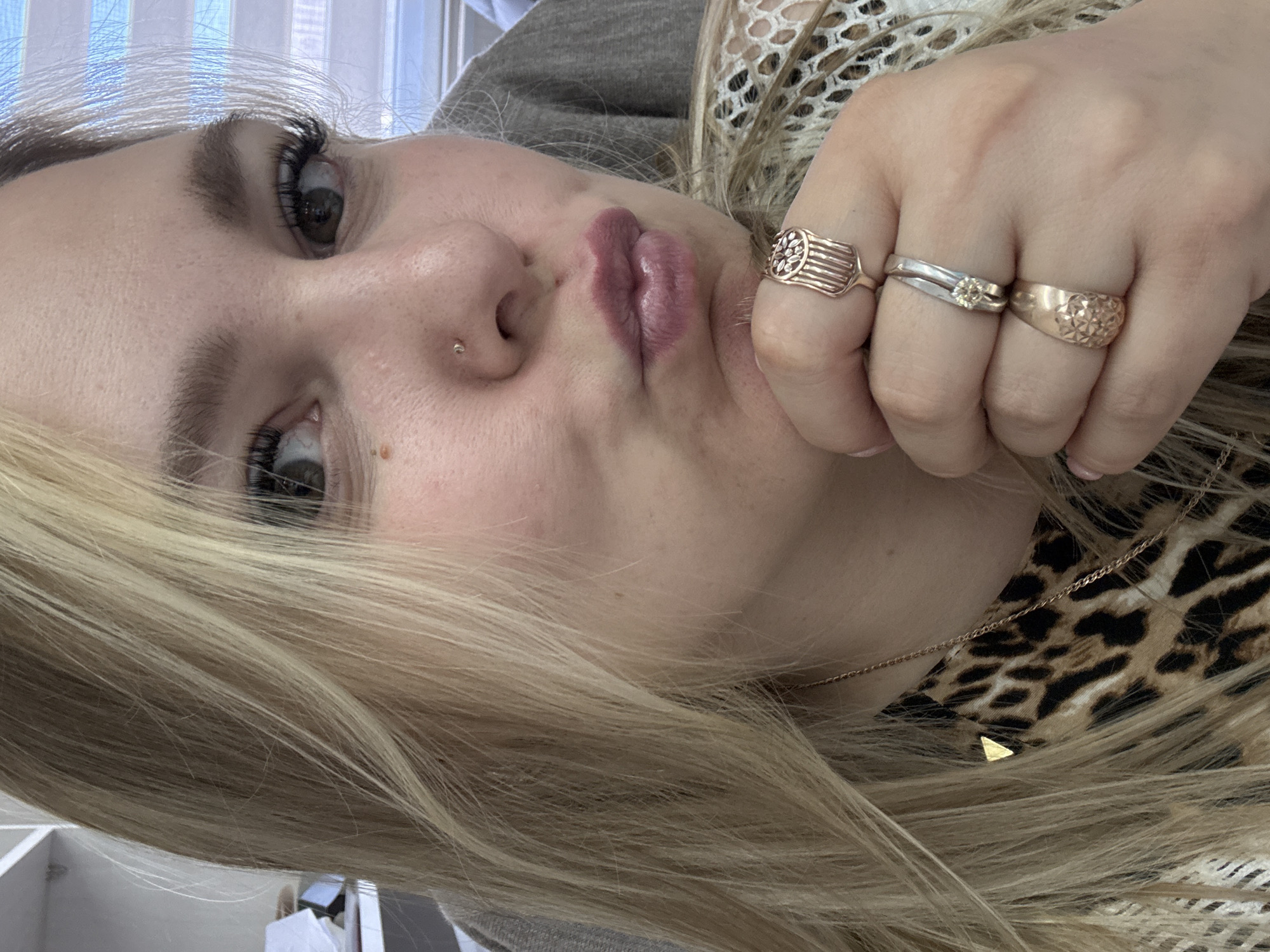 A close-up, angled portrait photograph depicts a young woman with blonde hair, fair skin, and noticeable makeup including dark eyeliner and mauve lipstick. She has a nose piercing on the left side of her nose. She is wearing three rings on one finger of her right hand: a ridged, gold-colored ring, a silver-colored ring with a small stone, and another gold-colored ring with a floral pattern. She also wears a thin gold necklace with a small triangle pendant. She appears to be wearing a gray top, a lace top, and a leopard print top. The background is softly out of focus and suggests a room with blinds. The location of Noviy, Russia is not relevant to the content of this image.