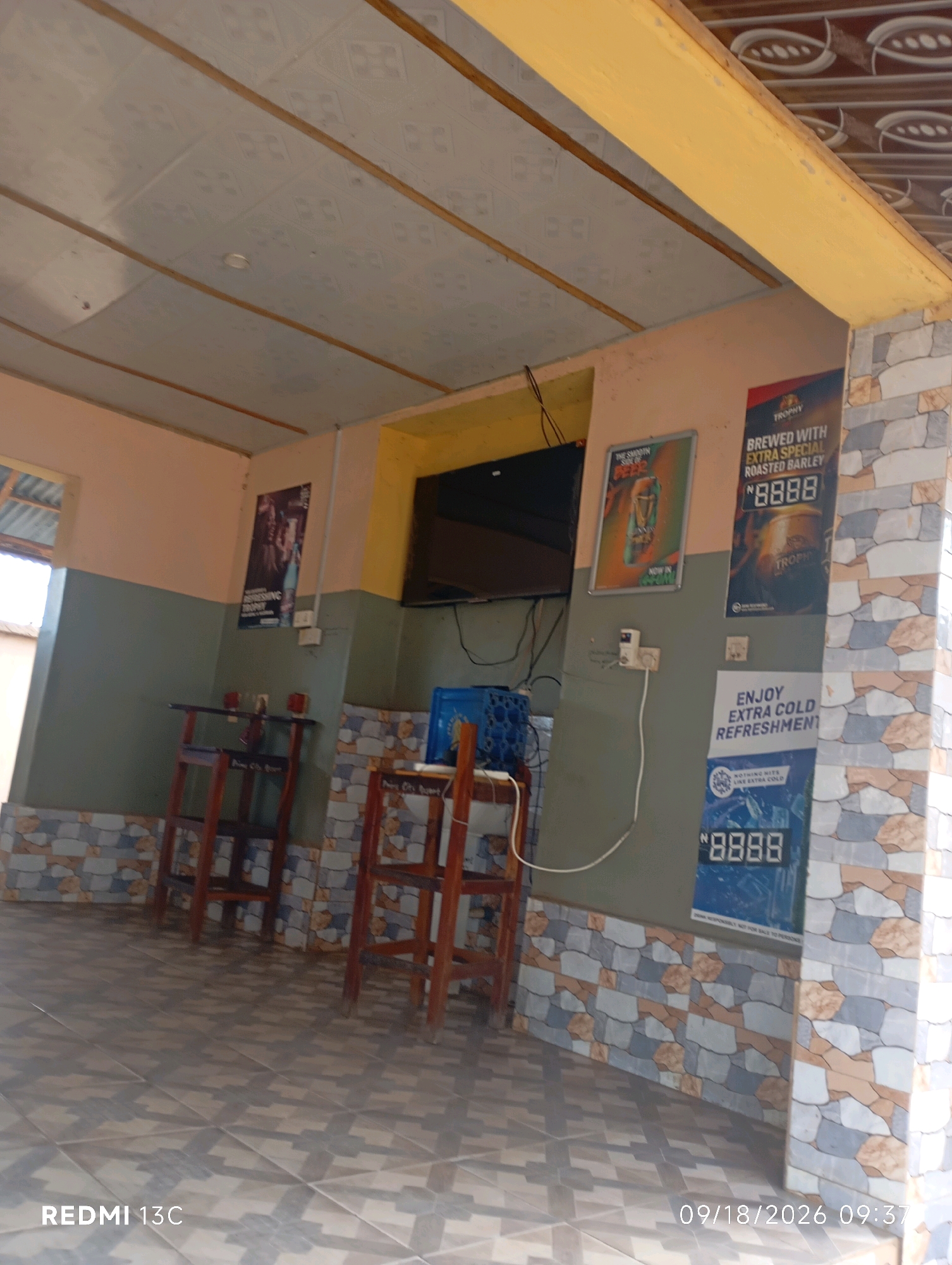 The image depicts an indoor scene, likely a bar or a local establishment, with a tiled floor and walls. The walls are painted in two tones, with a lighter color on top and a darker green below. A section of the wall is covered with decorative tiles featuring a mosaic pattern of light blue, gray, brown, and cream colors.
Several objects are visible:
* **Furniture:** Two wooden stools with high backs are present. One is in the foreground, and another is partially visible to its right.
* **Television:** A black flat-screen television is mounted on the wall.
* **Posters/Advertisements:** There are several posters on the wall. One shows a can of beer with the text "TROPHY" and "BREWED WITH EXTRA SPECIAL ROASTED BARLEY". Another poster features people and the word "REFRESHING". A third poster, also blue, promotes "ENJOY EXTRA COLD REFRESHMENT" and shows a price of "N 8888".
* **Electrical Outlets:** Electrical outlets and wiring are visible on the wall.
* **Blue Crate:** A blue plastic crate is placed on the floor near the television.
* **Ceiling:** The ceiling is paneled with what appears to be acoustic tiles and wooden beams.
The overall impression is of a casually decorated interior, possibly a local spot for drinks. The presence of the posters advertising alcoholic beverages and the stools suggest a place where patrons would sit and consume drinks. The tiling on the walls and floor provides a decorative and potentially easy-to-clean surface. The date and time stamp "09/18/2026 09:37" and the text "REDMI 13C" suggest this image was taken with a Redmi 13C phone on that date and time. There are no people visible in the image. The location is identified as the city of Shendam, Nigeria.