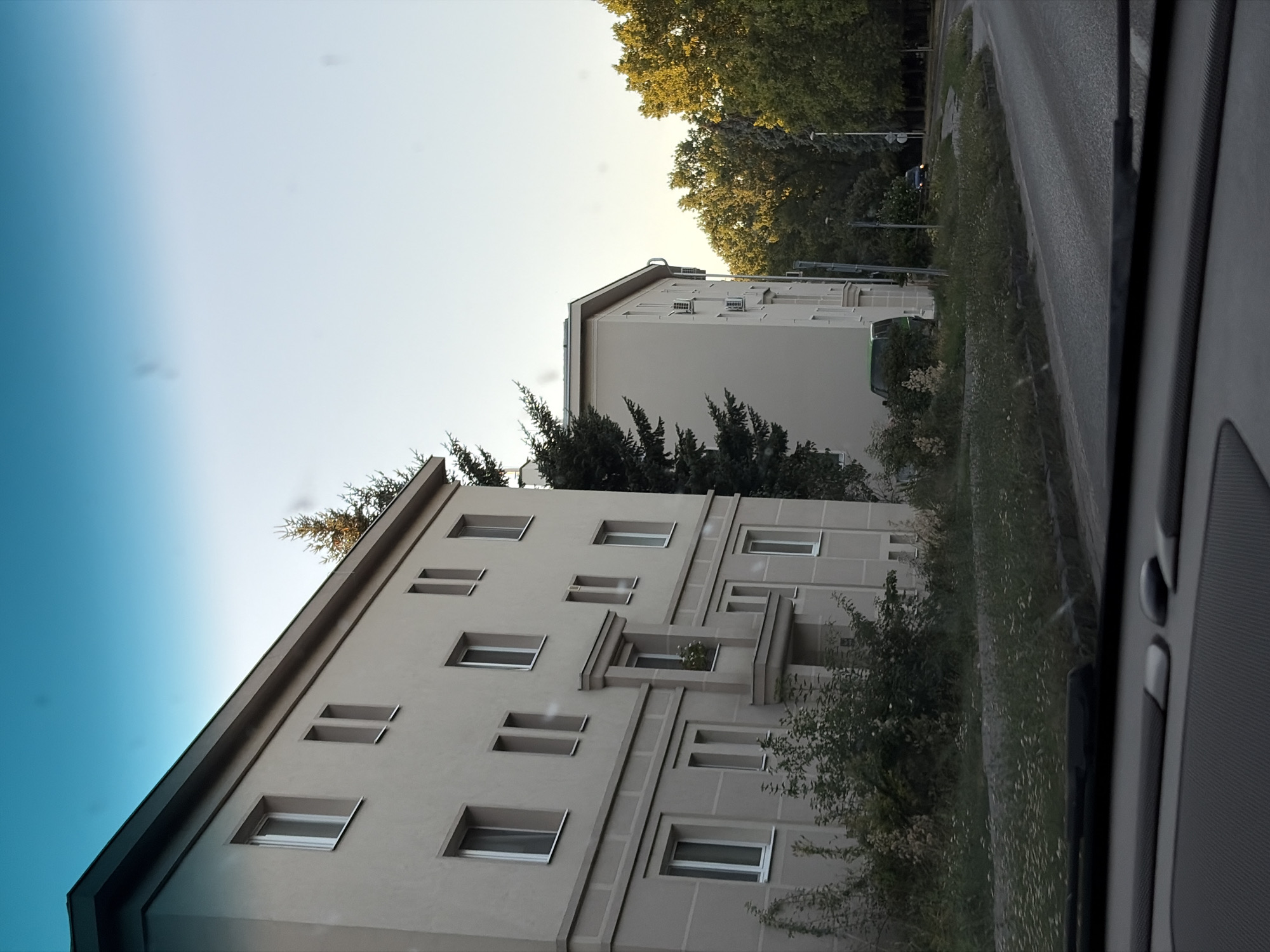 The image captures a street view in the city of Nitra, Slovakia, likely in a residential area. The foreground shows the interior of a vehicle, with a dark dashboard and windshield wipers visible. The perspective is from inside the car, looking outwards.
The main subjects are two multi-story apartment buildings constructed in a functionalist architectural style, characterized by simple geometric shapes and plain facades. The building on the left is larger and closer, with multiple windows visible on its beige-colored exterior. The building on the right is slightly further away and partially obscured by trees. Both buildings appear to be residential, with small balconies or projecting window elements.
The environment consists of a paved road curving through the scene, bordered by grassy areas with scattered wildflowers. Dense foliage and trees, some with golden autumn leaves, line the background, suggesting a natural or park-like setting adjacent to the urban development. A green van is parked near the building on the right, and another dark-colored vehicle is visible further down the road. The sky is a clear, light blue, indicating daytime. The overall impression is one of a quiet, somewhat overcast residential street in a European city.