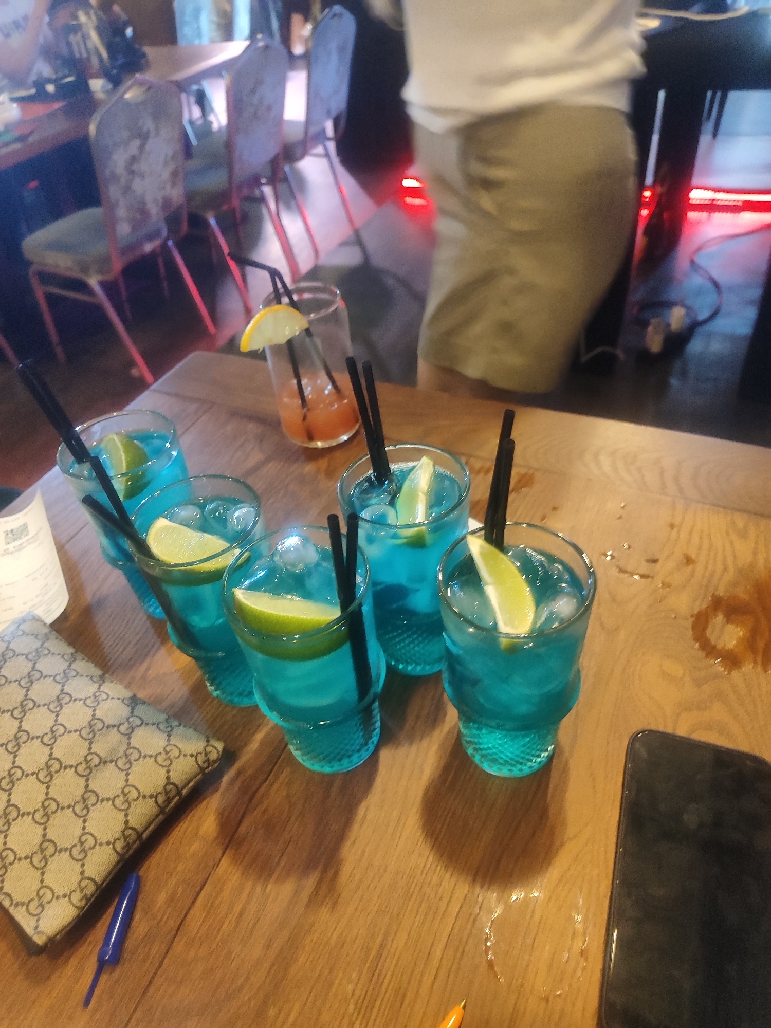 The image shows a wooden table with several glasses of blue liquid, garnished with lime wedges and black straws. There is also a glass with orange liquid and a lemon wedge in the background. A wallet with a logo pattern and a blue pen are on the table in the foreground. A smartphone is visible on the right side of the table. In the background, several chairs and tables are visible, suggesting an indoor establishment such as a bar or restaurant. The lighting in the scene is dim with some red accents.